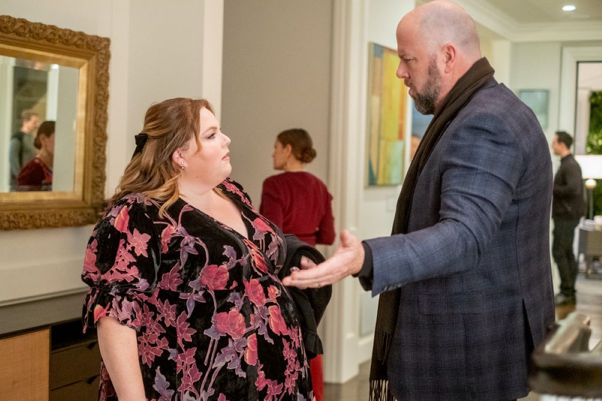 'This Is Us' stars Chrissy Metz and Chris Sullivan, in character as Kate and Toby, share a scene. Kate wears a black dress with red and purple flowers. Toby wears a blue plaid coat and brown scarf.