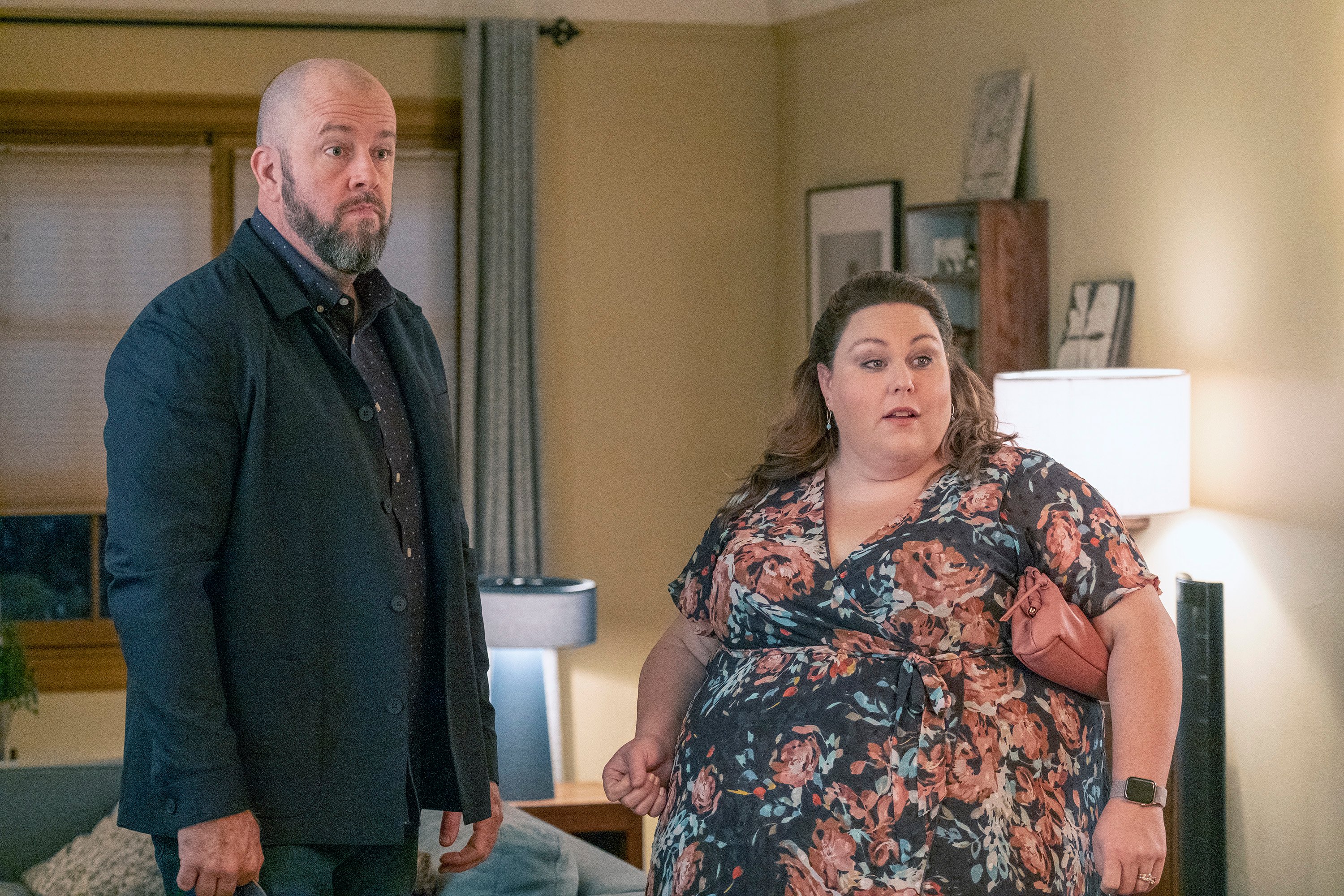 Chris Sullivan and Chrissy Metz, who play Toby and Kate in 'This Is Us' Season 6 Episode 9, share a scene. Toby wears a black jacket over a dark blue button-up shirt with white polka dots and jeans. Kate wears a dark blue short-sleeved dress with dark pink roses on it.