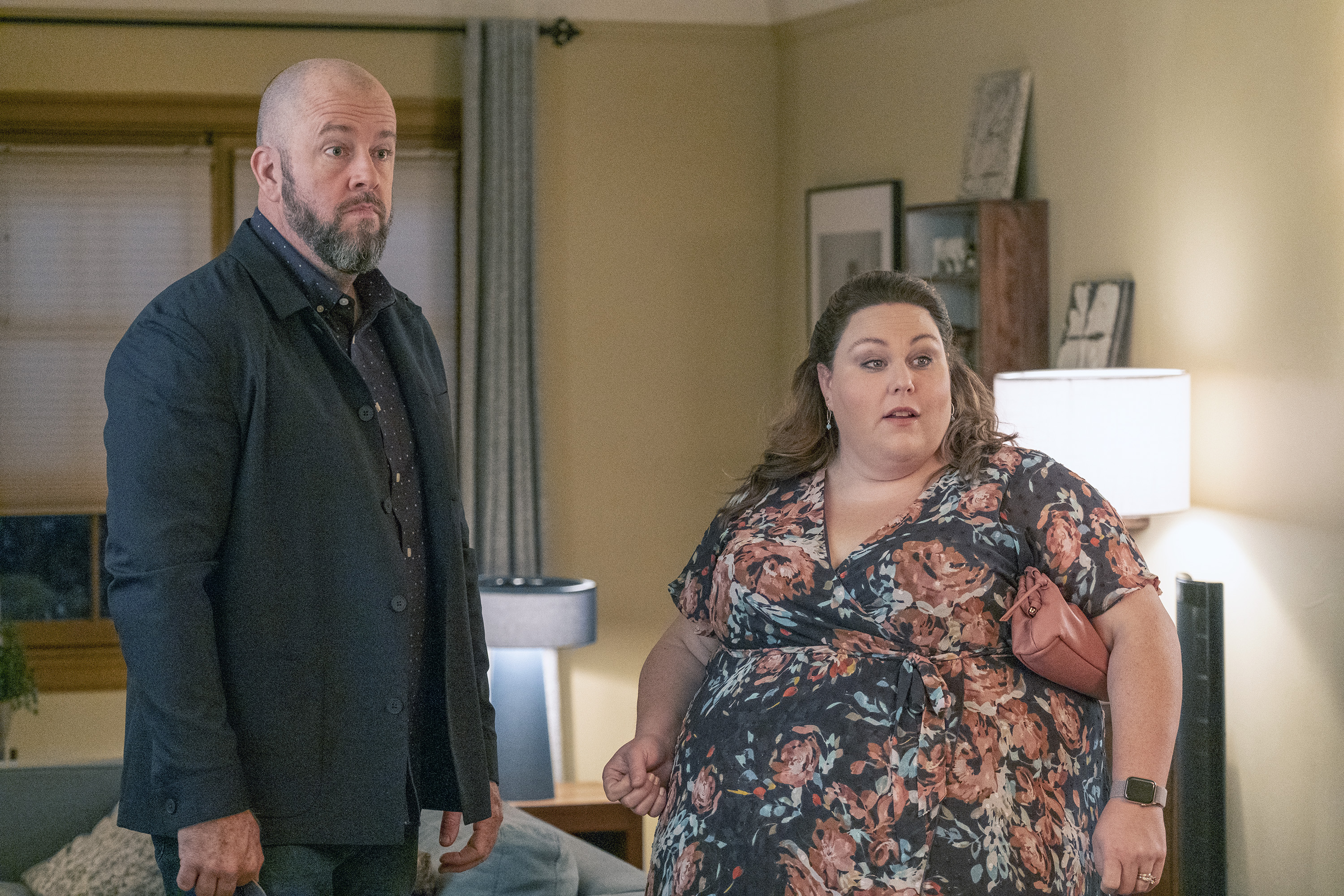 Chris Sullivan and Chrissy Metz, who play Toby and Kate in 'This Is Us' Season 6 Episode 9, share a scene. Toby wears a black jacket over a dark blue button-up shirt with white polka dots and jeans. Kate wears a dark blue short-sleeved dress with dark pink roses on it.