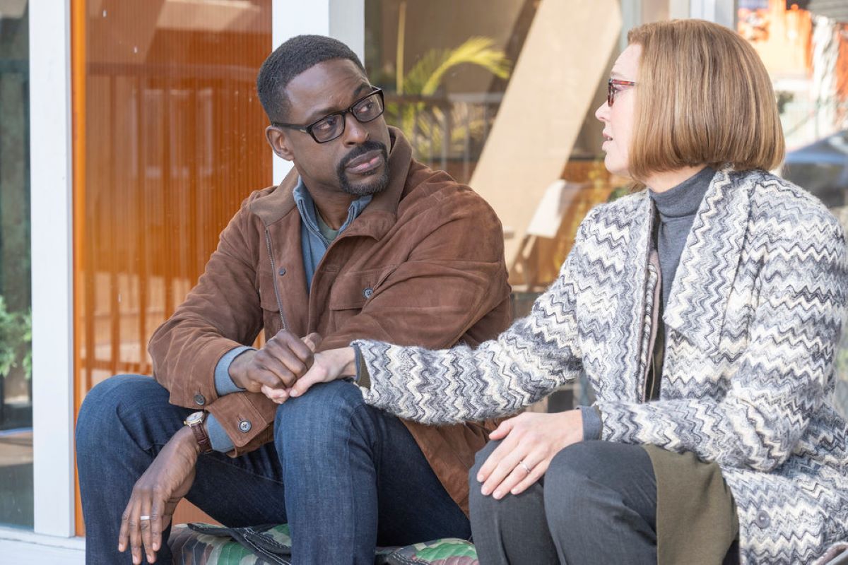 'This Is Us' Season 6 Episode 10 stars Sterling K. Brown and Mandy Moore, in character as Randall and Rebecca, share a scene and hold hands. Randall wears a brown jacket over a blue sweater and jeans. Rebecca wears a white and blue zig-zag patterned coat over a blue turtleneck and dark gray pants.