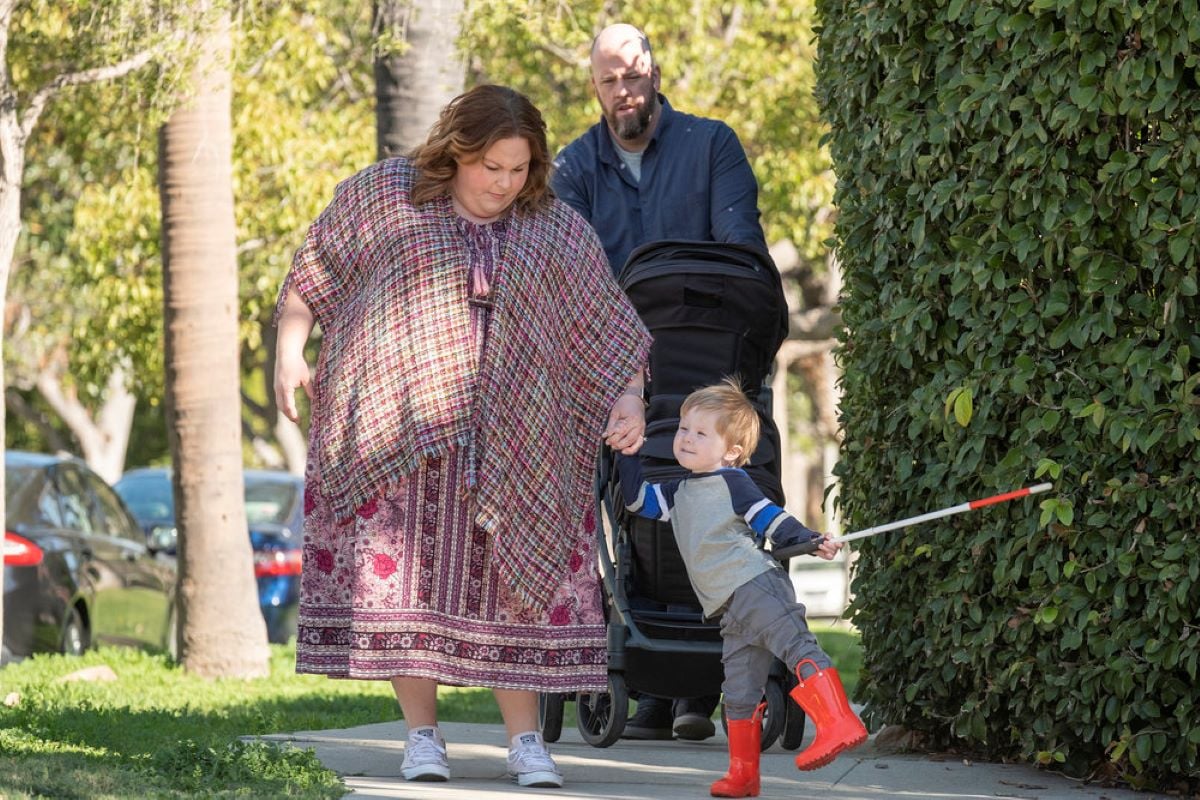 'This Is Us' Season 6 Episode 11 stars Chrissy Metz, Chris Sullivan, and Jonathan Kincaid, in character as Kate, Toby, and Jack, share a scene while walking on the sidewalk. Kate wears a pink, purple, and white floral dress and a multi-colored shawl. Toby wears a blue button-up shirt and pushes a stroller. Jack, who is holding Kate's hand in one hand and his white cane in the other, wears a blue and white shirt, gray pants, and red rain boots.