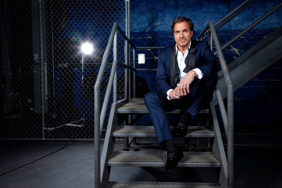 'The Bold and the Beautiful' actor Thorsten Kaye wearing a navy blue suit; sits on a staircase in front of a chainlink fence.