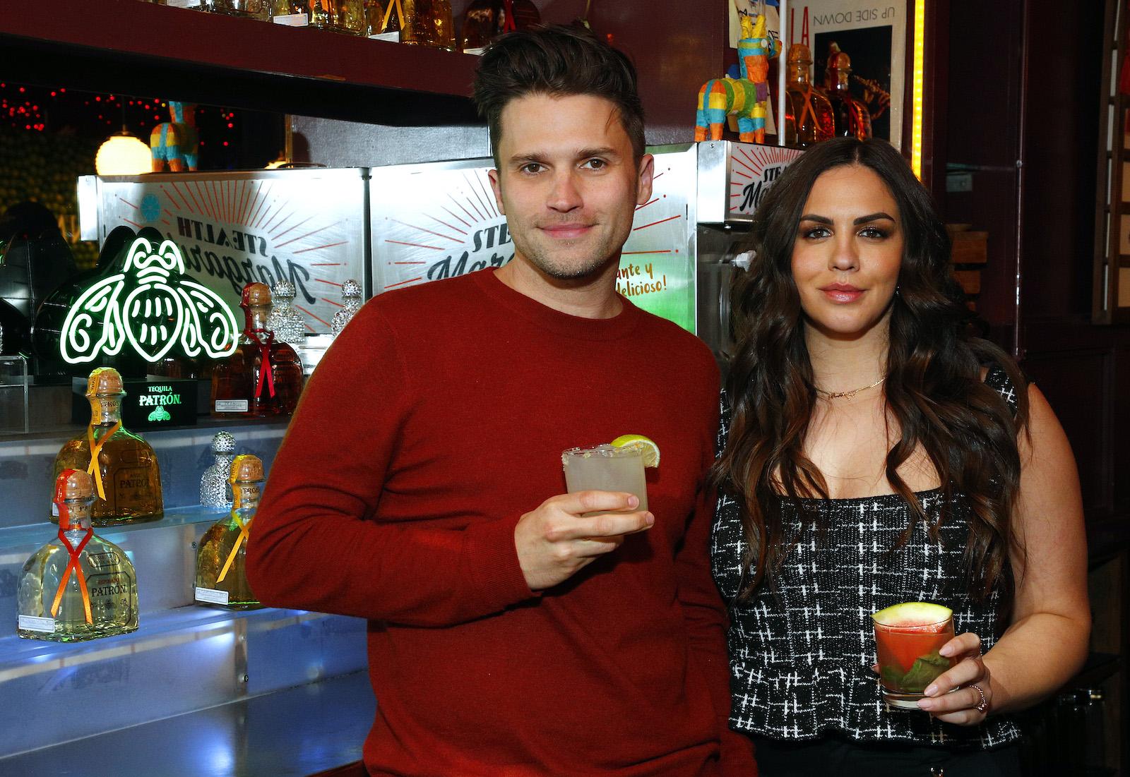Tom Schwartz and Katie Maloney-Schwartz from 'Vanderpump Rules' hold cocktails and stand by a bar smiling 