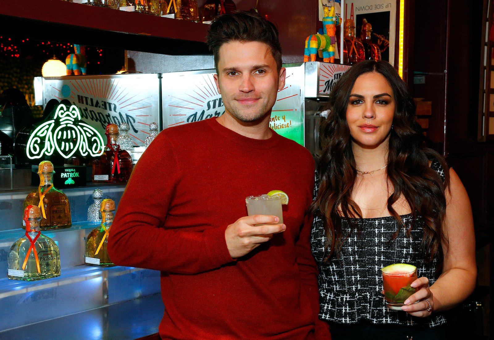 Tom Schwartz and Katie Maloney-Schwartz from 'Vanderpump Rules' hold cocktails and stand by a bar smiling 