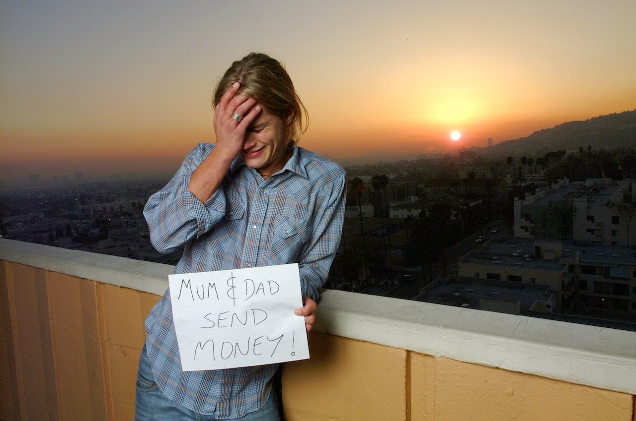 Travis Fimmel in 2002 when he was a new Calvin Klein model, standing on a balcony in front of a sunset holding a sign that reads 'Mum & Dad Send Money'