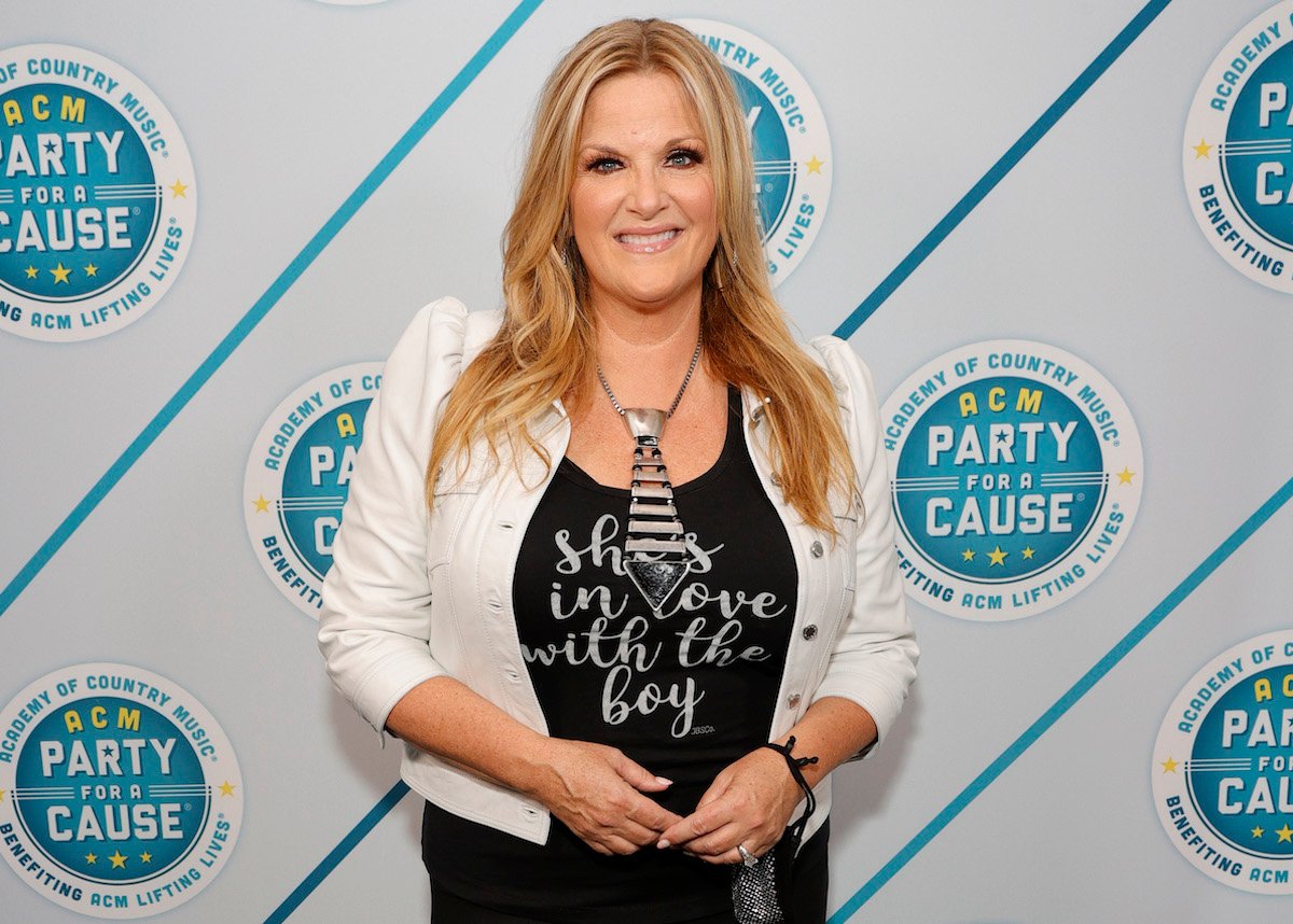 Trisha Yearwood smiles wearing a white jacket over a gray t-shirt reading, 'She's in love with the boy.'