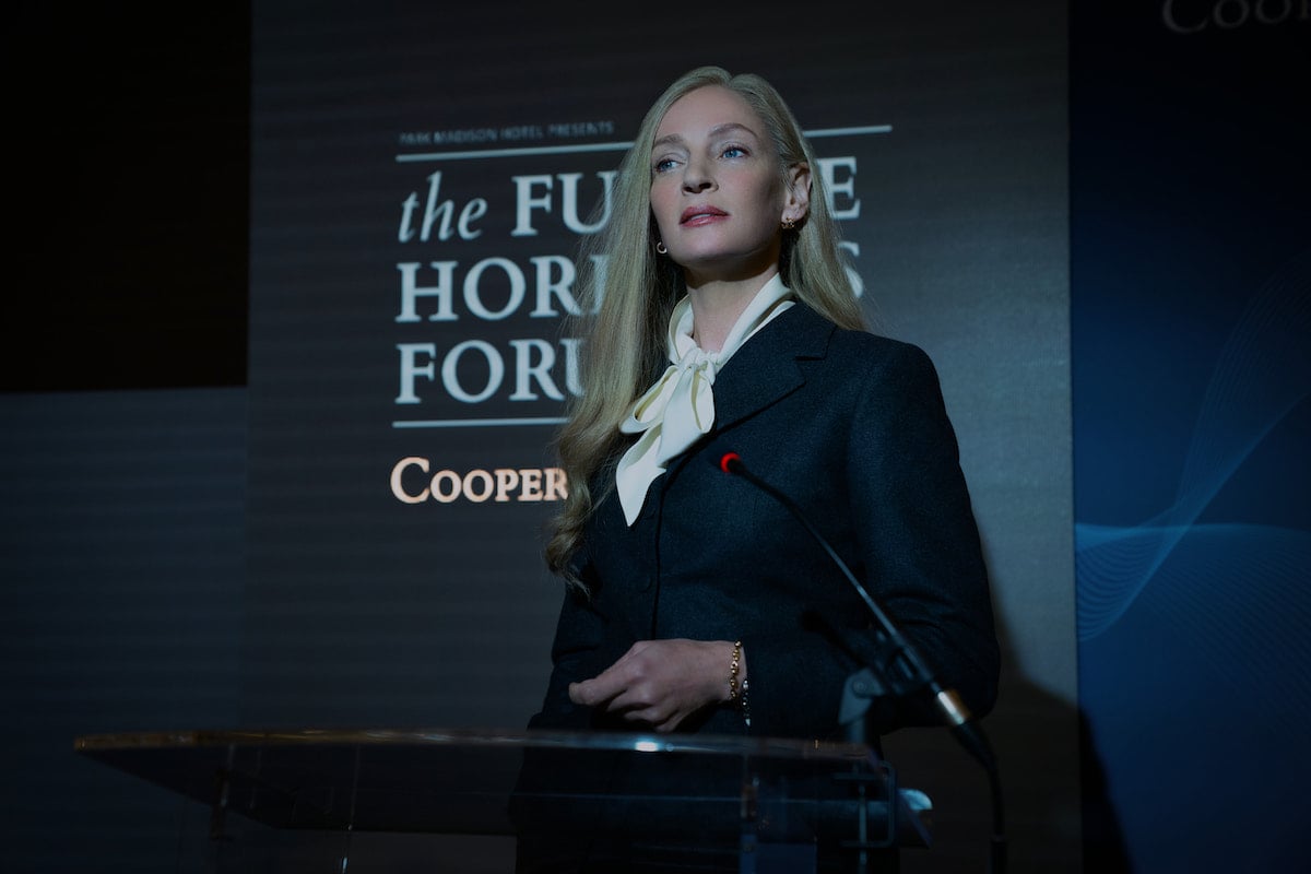 Uma Thurman stands at a podium wearing a black and white suit in 'Suspicion' Season 1 Episode 8: 'Unmasked'