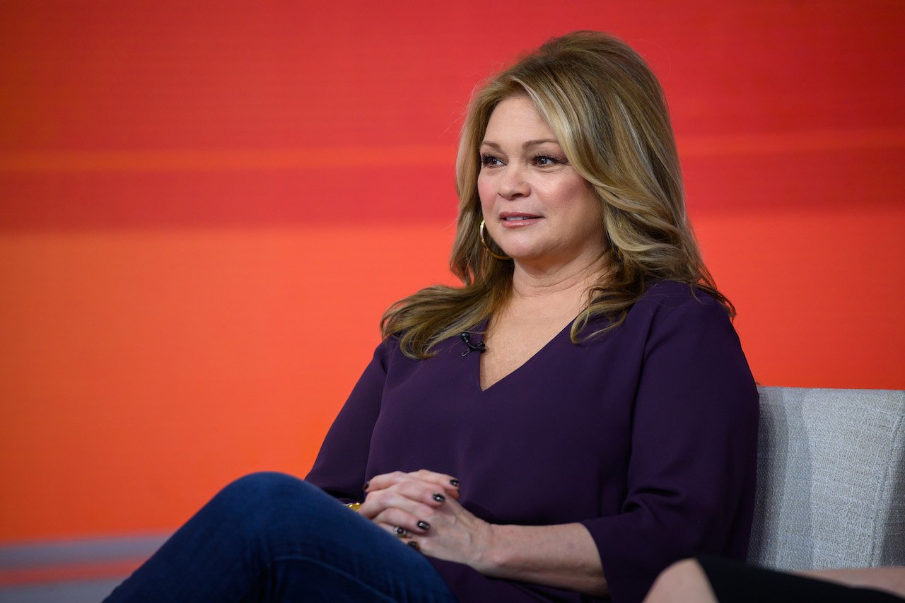 Valerie Bertinelli in a purple top, sitting with her legs crossed and her hands clasped together