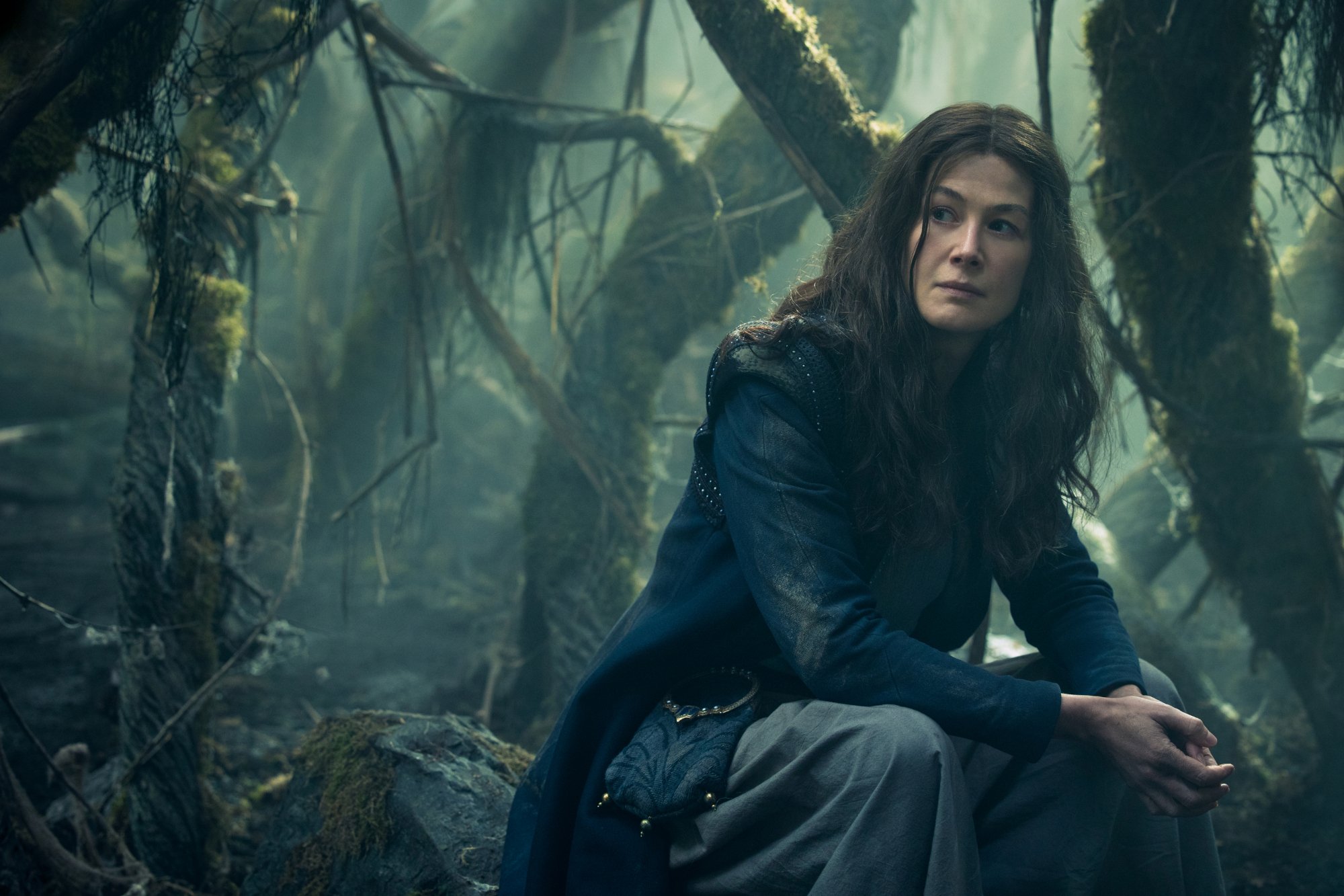 Rosamund Pike as Moiraine Damodred in 'The Wheel of Time' TV series. She's kneeling in a dark wood and looking at something off-screen.