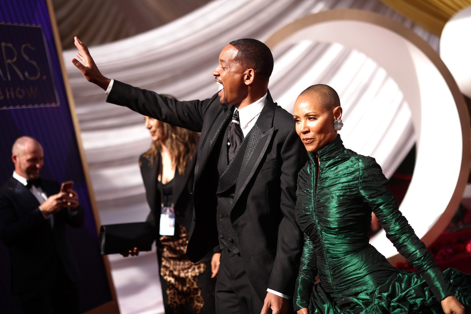 Will Smith and Jada Pinkett Smith on the red carpet at the Oscars 2022. Will Smith is waving with his mouth open excitedly