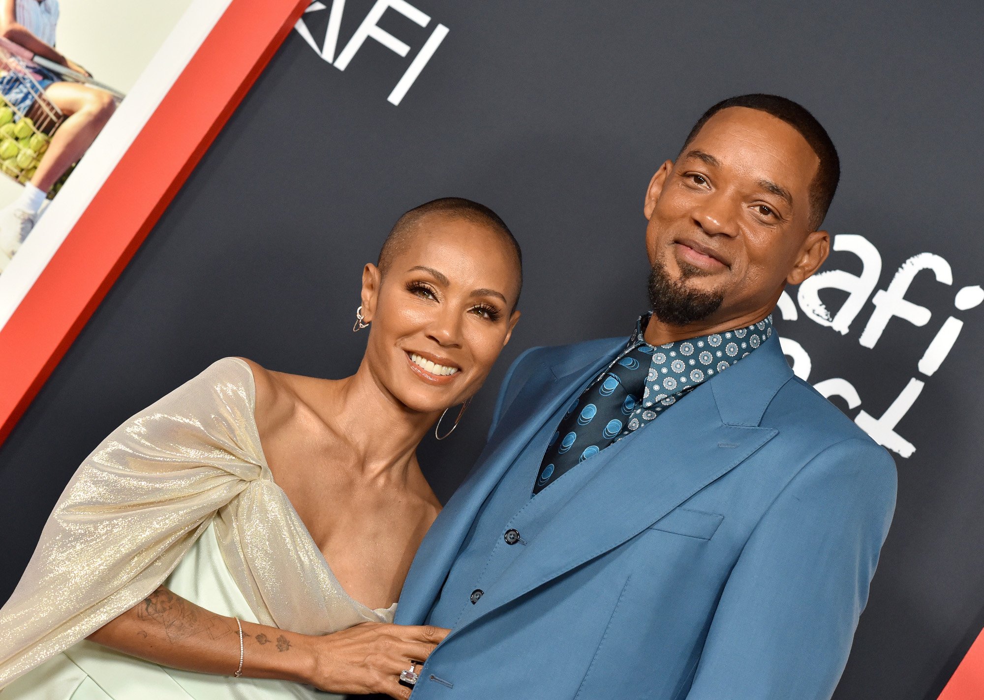 Jada Pinkett Smith and Will Smith. She's wearing a biege, off-the-shoulder dress. He's wearing a medium blue suit.