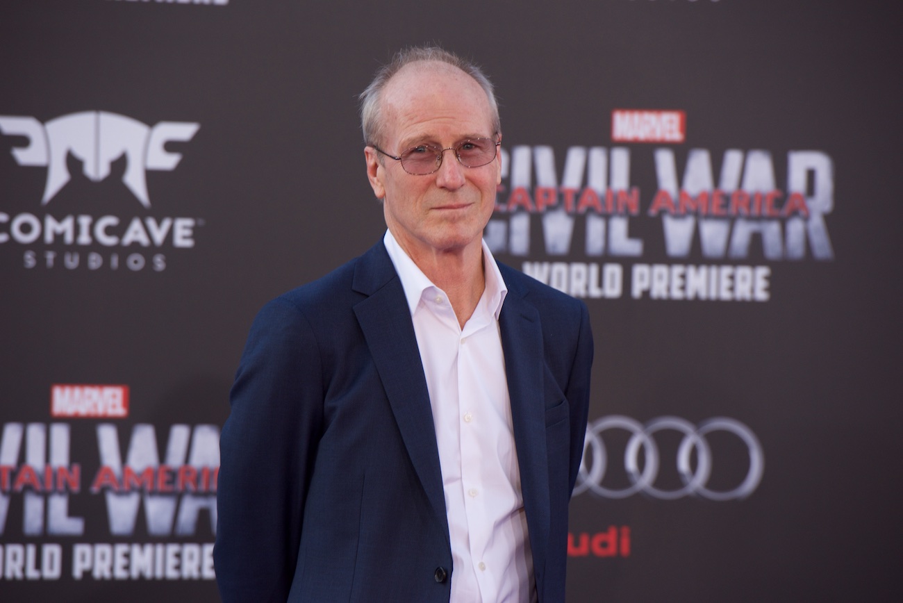 William Hurt wearing a white shirt and dark jacket, standing in front of a dark background with white text