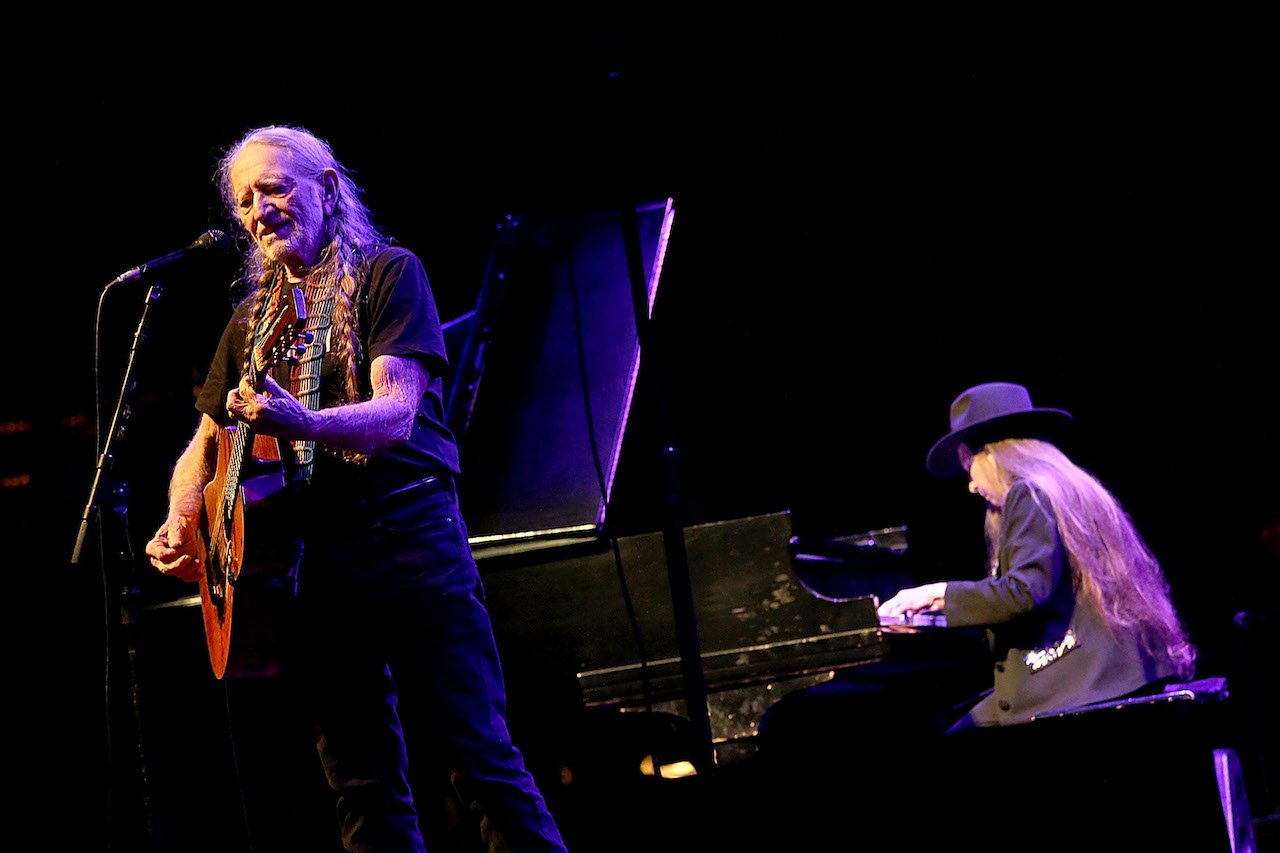 Willie Nelson stands in front of a microphone, holding a guitar and Bobbie Nelson sits at the piano behind him