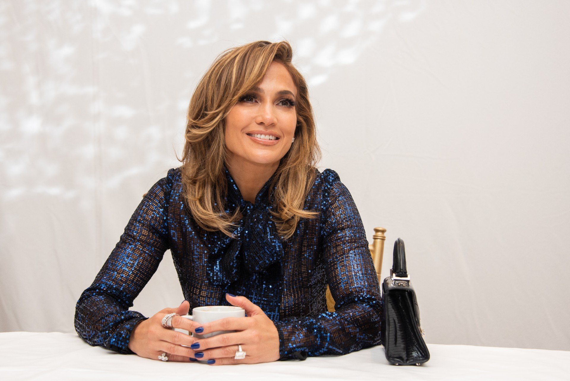 Jennifer Lopez attends a Hustlers press conference while wearing a blue, sparkly outfit and holding a coffee cup. 