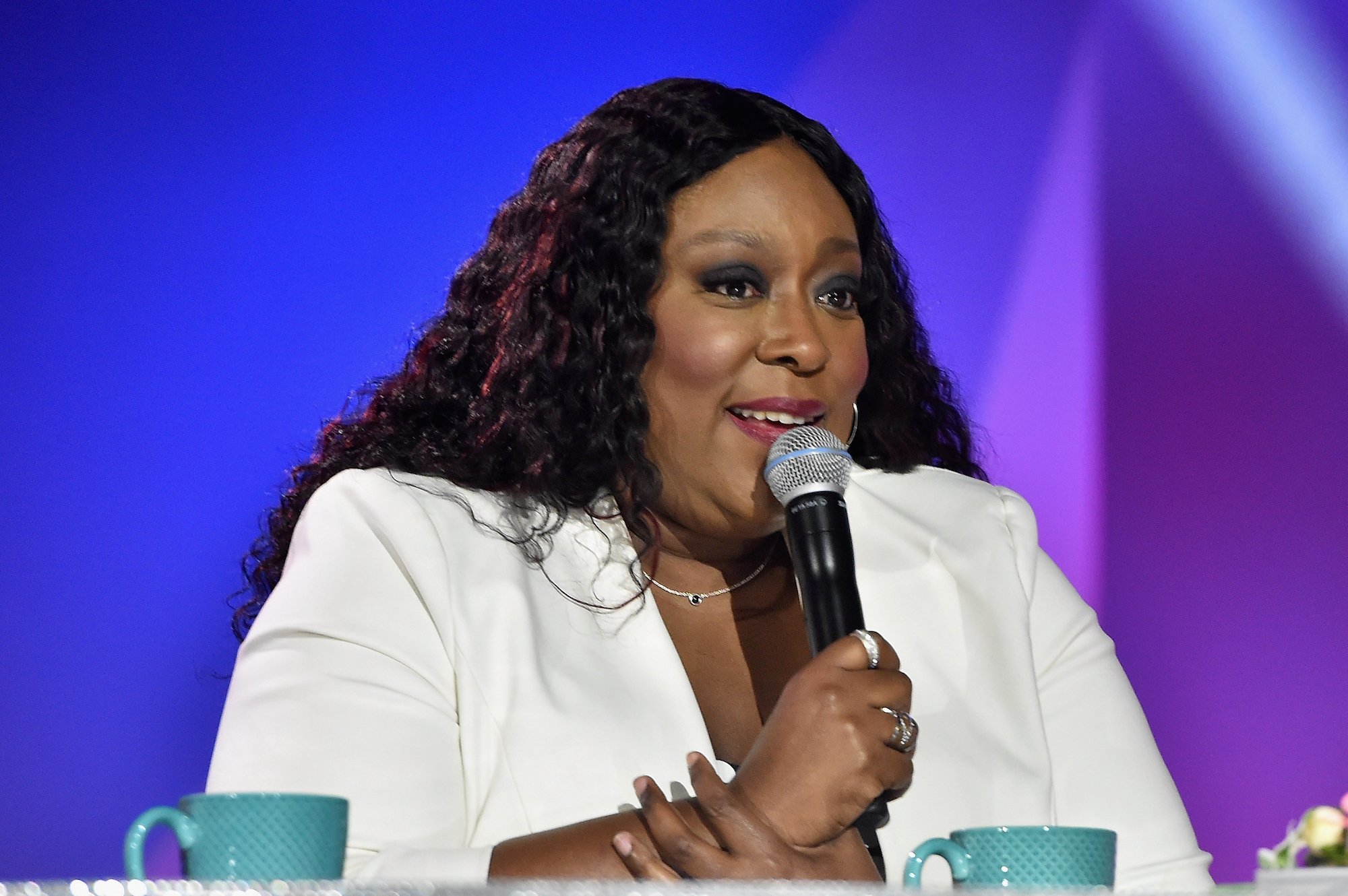 Loni Love holding a microphone and wearing a white blazer