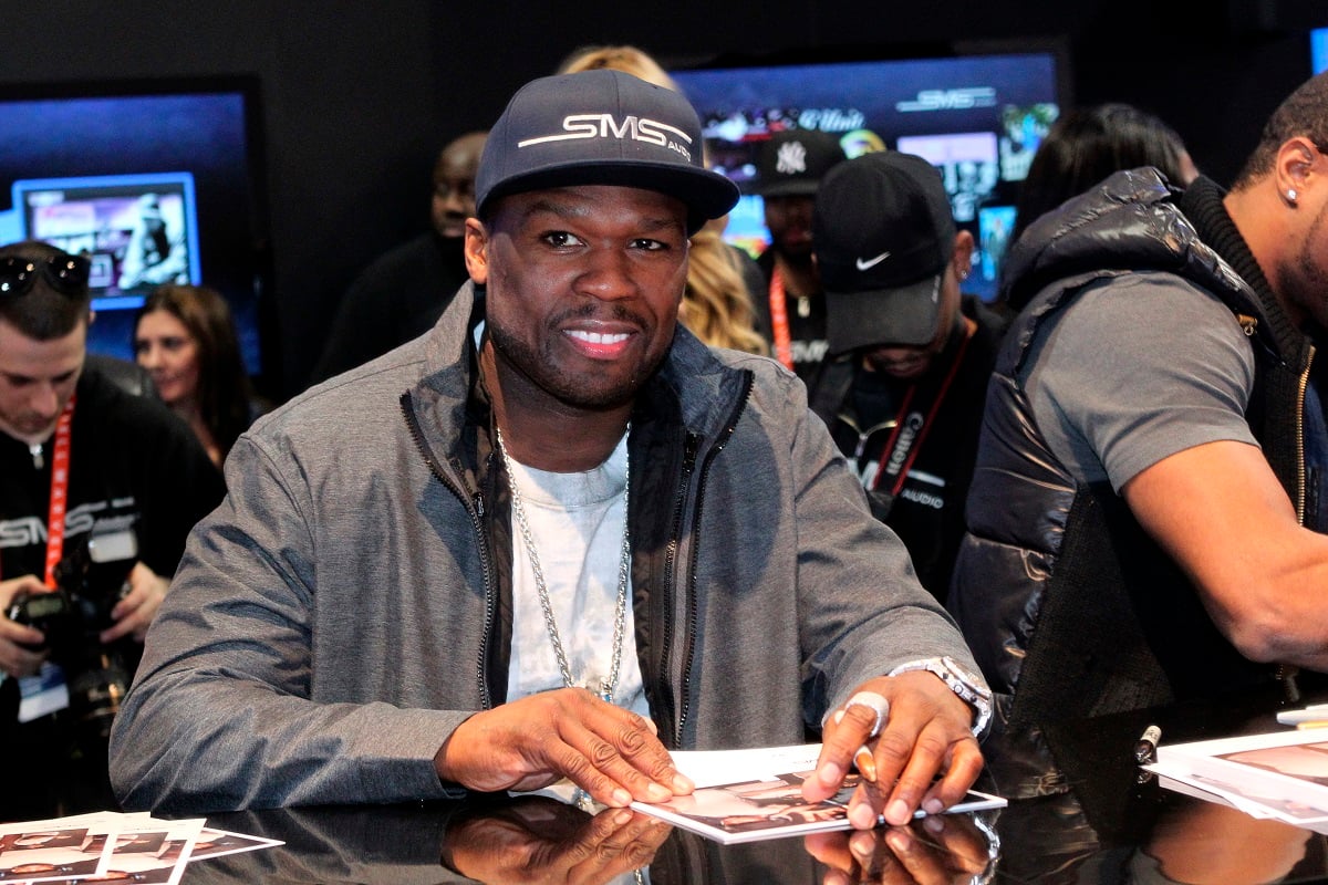 50 Cent smiling while wearing a grey jacket and a cap.