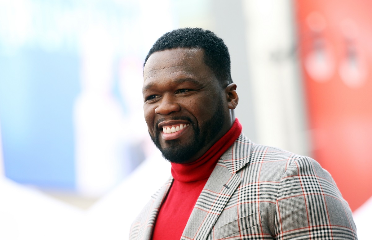 50 Cent smiling while wearing a red turtleneck and a suit.