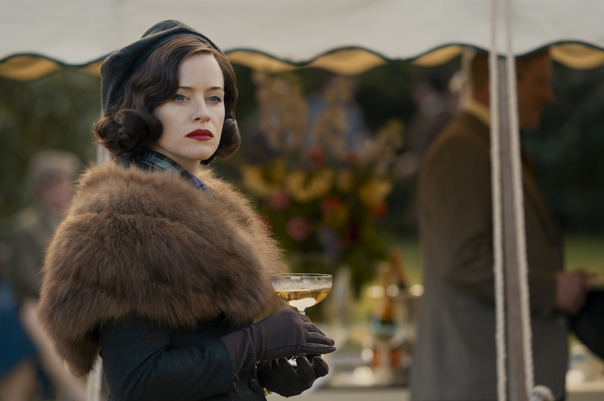 Claire Foy wearing a hat and a fur stole and holding a champagne coupe in 'A Very British Scandal'