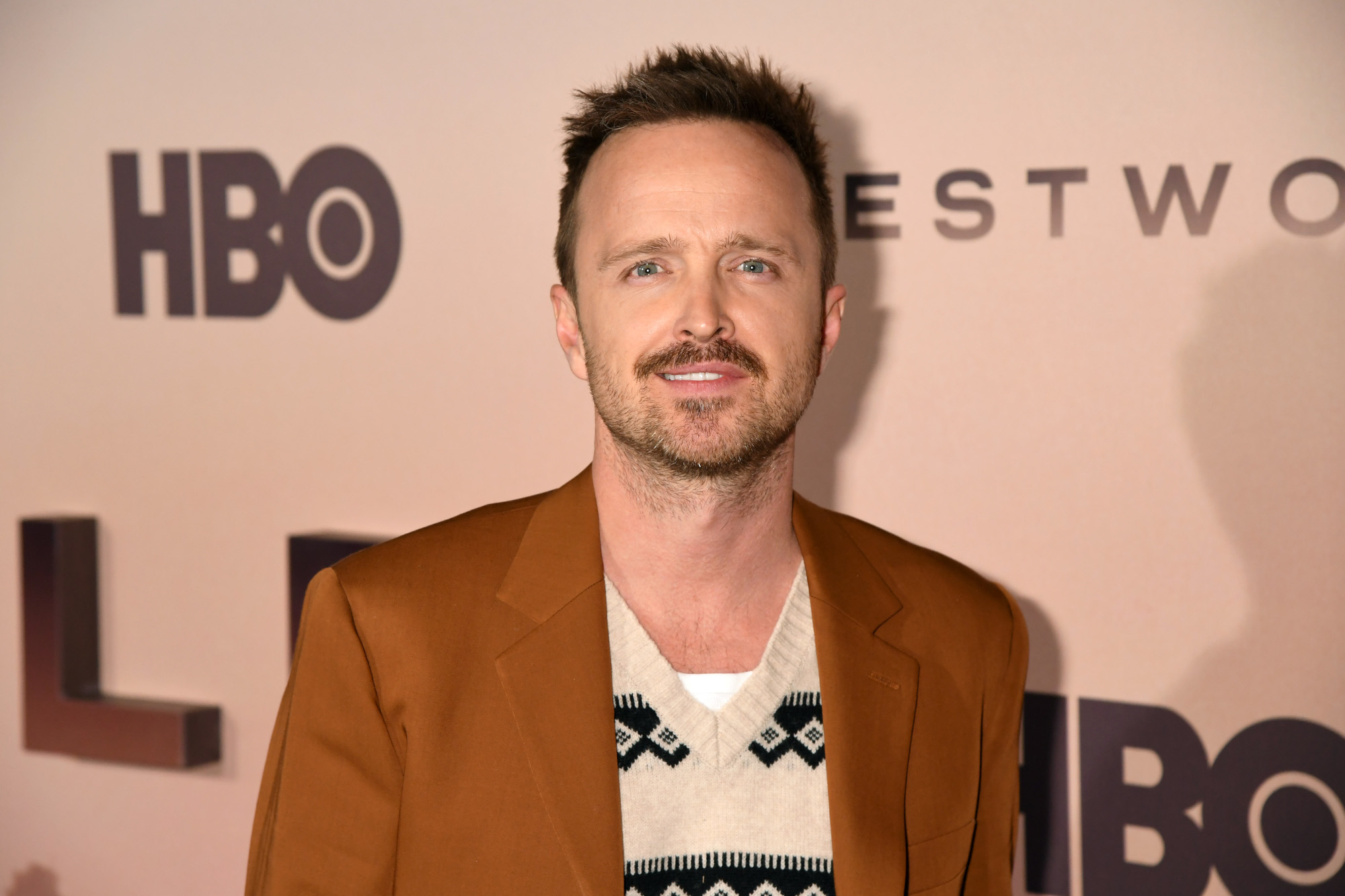 'Breaking Bad' and 'Better Call Saul' star Aaron Paul. He's wearing a beige and black sweater with a brown blazer. He's standing in front of a wall promoting HBO's 'Westworld' and smiling.