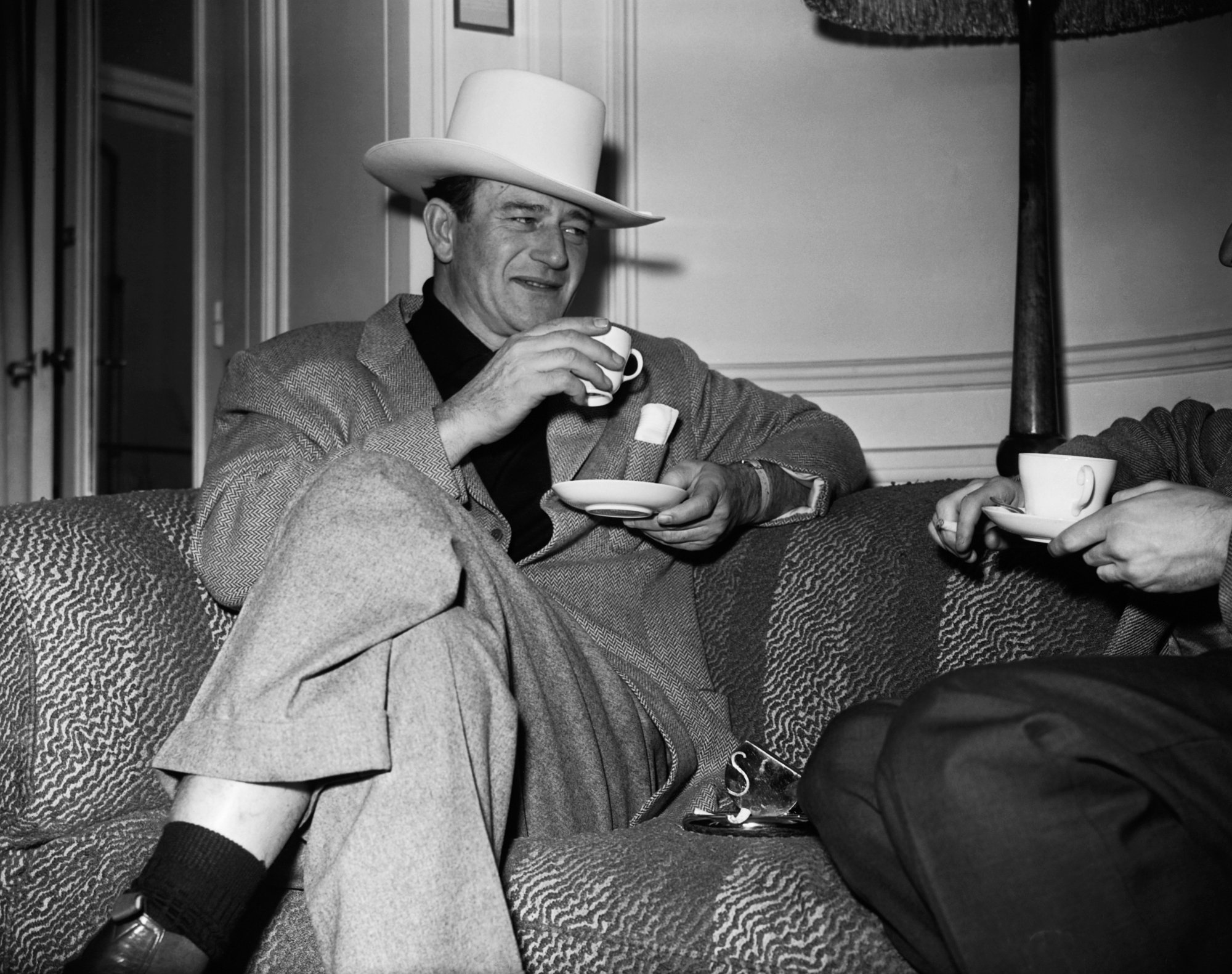 Actor John Wayne, who believed himself to be a liberal, wearing a suit and drinking from a tea cup