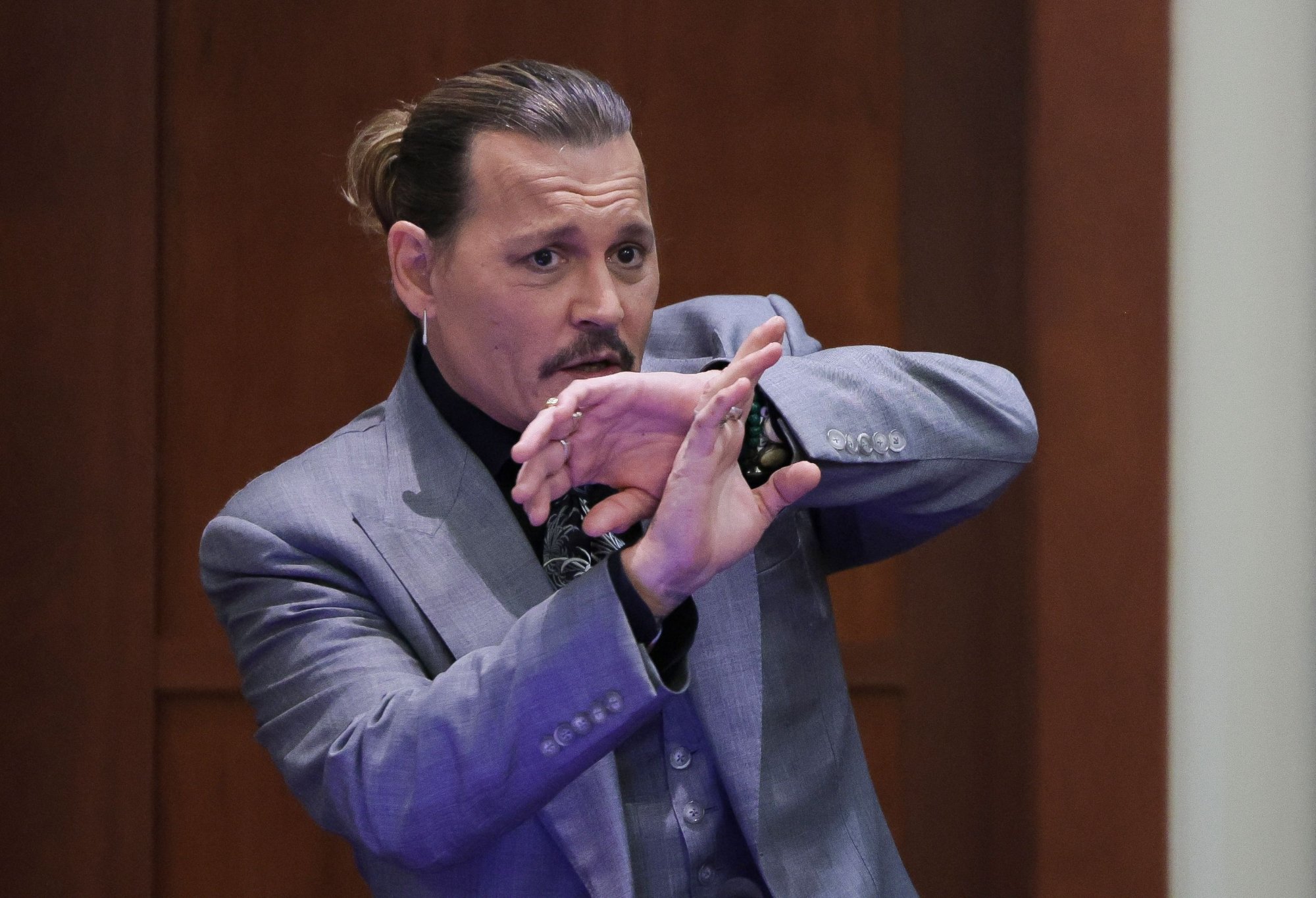 Actor Johnny Depp testifying in trial against ex-wife, Amber Heard, who he alleged pooped in his bed as revenge wearing a grey suit and holding his hands up in a defensive pose