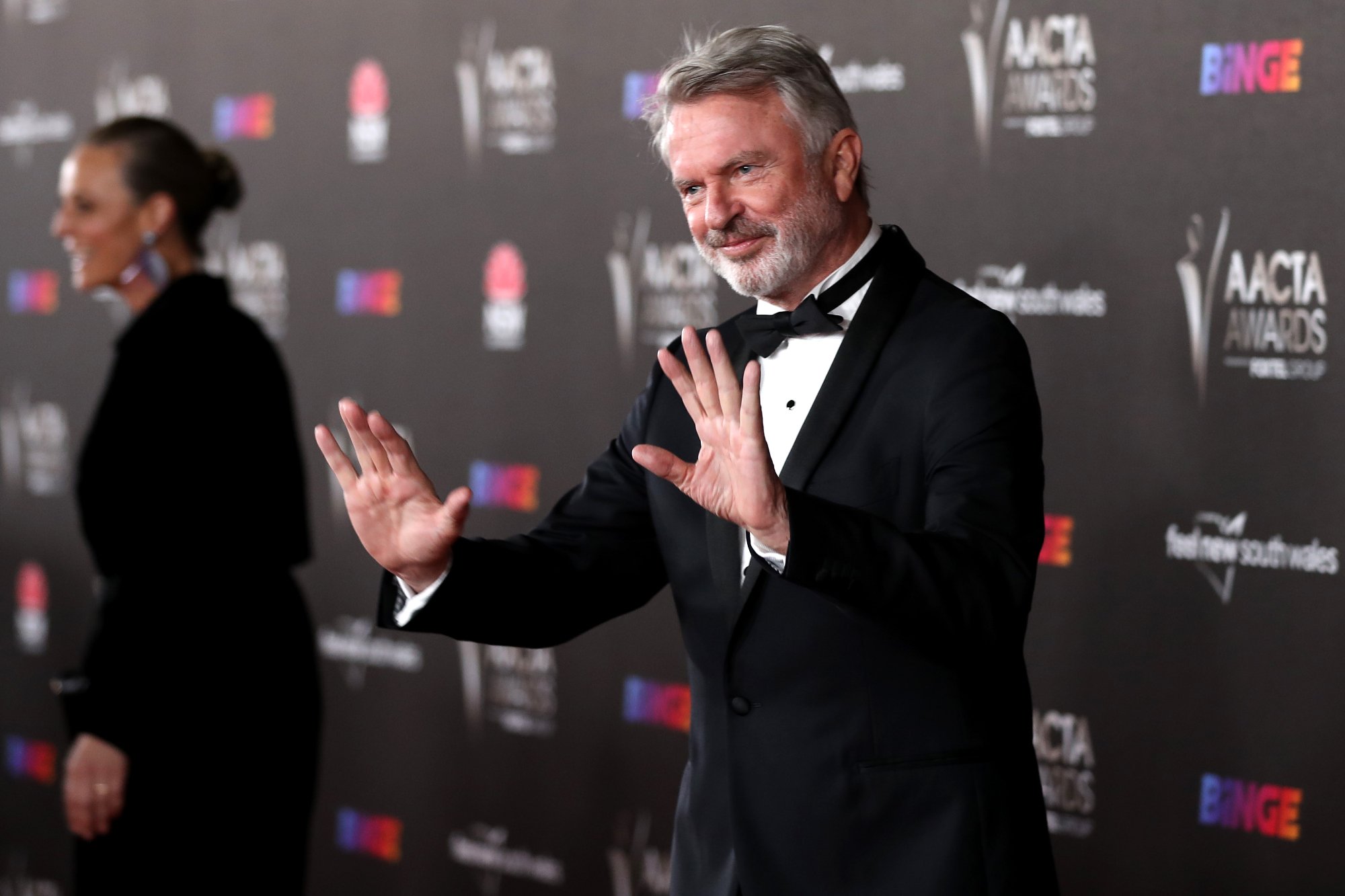 Actor Sam Neill wearing a tux holding his hands up at the 2021 AACTA Awards in front of a step and repeat