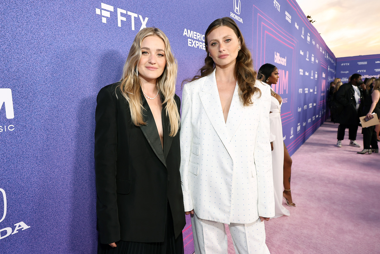 Amanda Michalka wearing a black oversized suit and Aly Michalka wearing a white oversized suit, both standing in front of a purple background