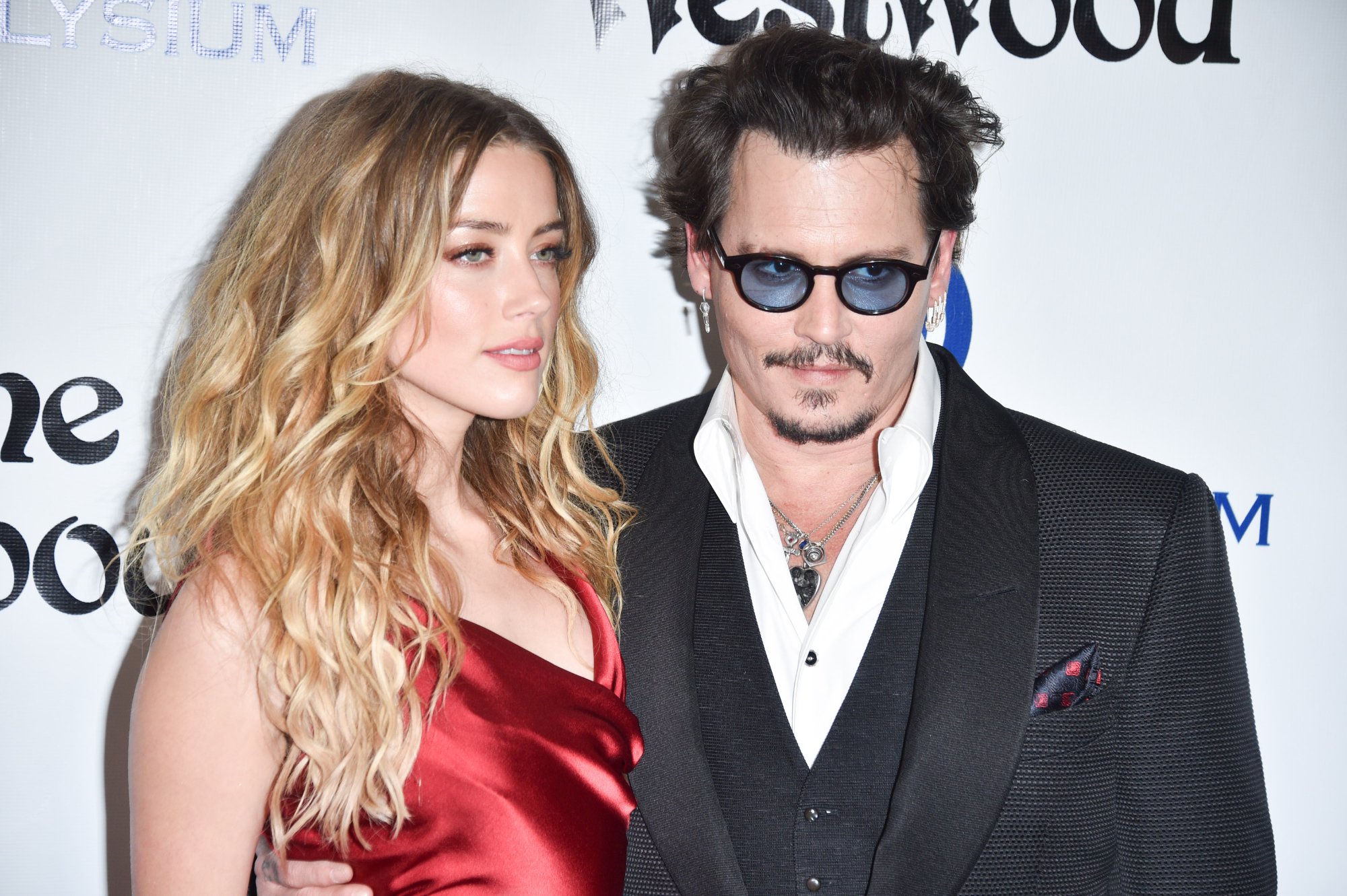 Amber Heard and Johnny Depp, who are in a court trial, standing in front of a white step and repeat with Heard in a red dress and Depp in a suit