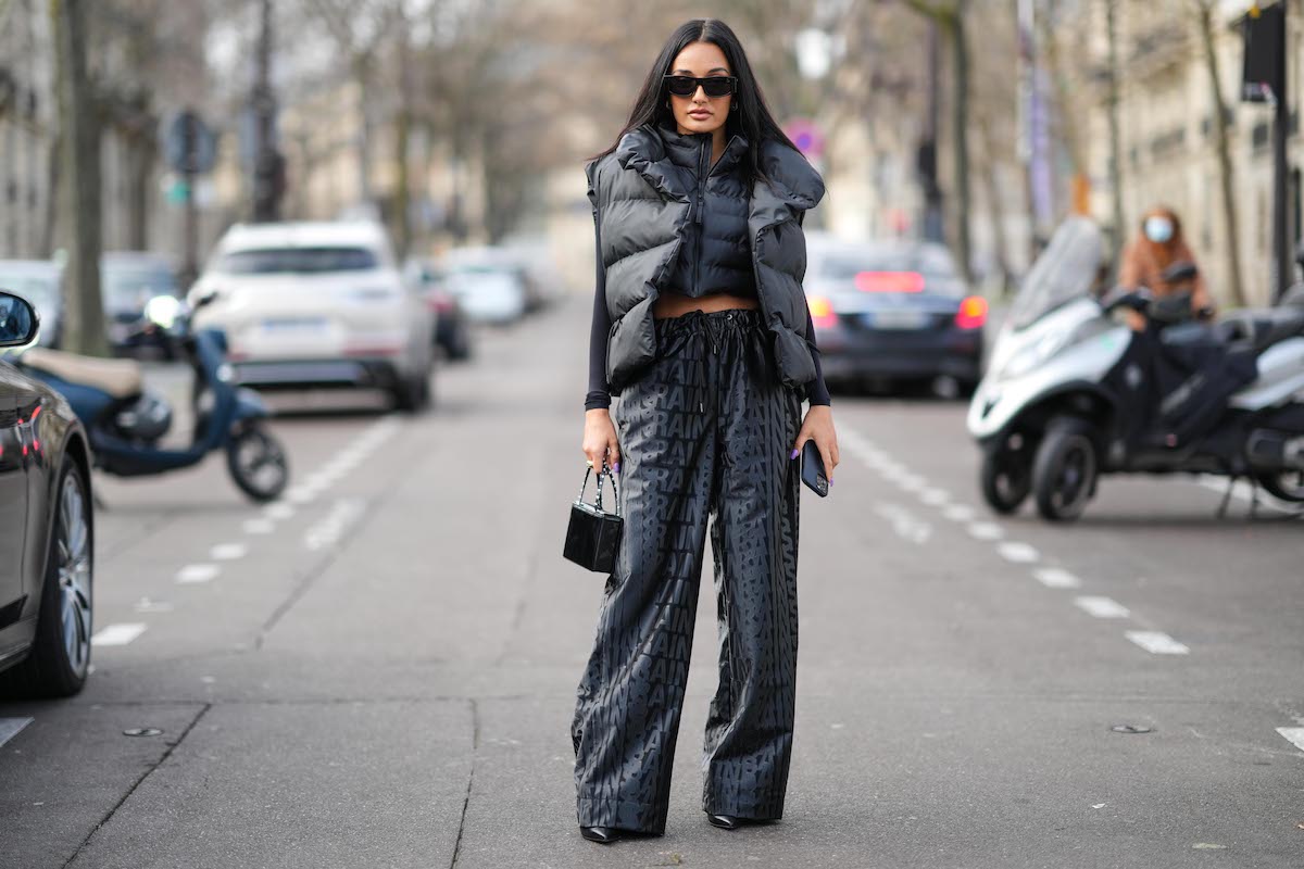 Fashion designer Amina Muaddi wears sunglasses, an oversized sleeveless gray winter puffer jacket, a small cropped dark gray puffer jacket, a black top, black and gray monogram/logo printed flared pants from Rains, a black leather bejeweled mini bag, outside Rains during Paris Fashion Week 2022-2023