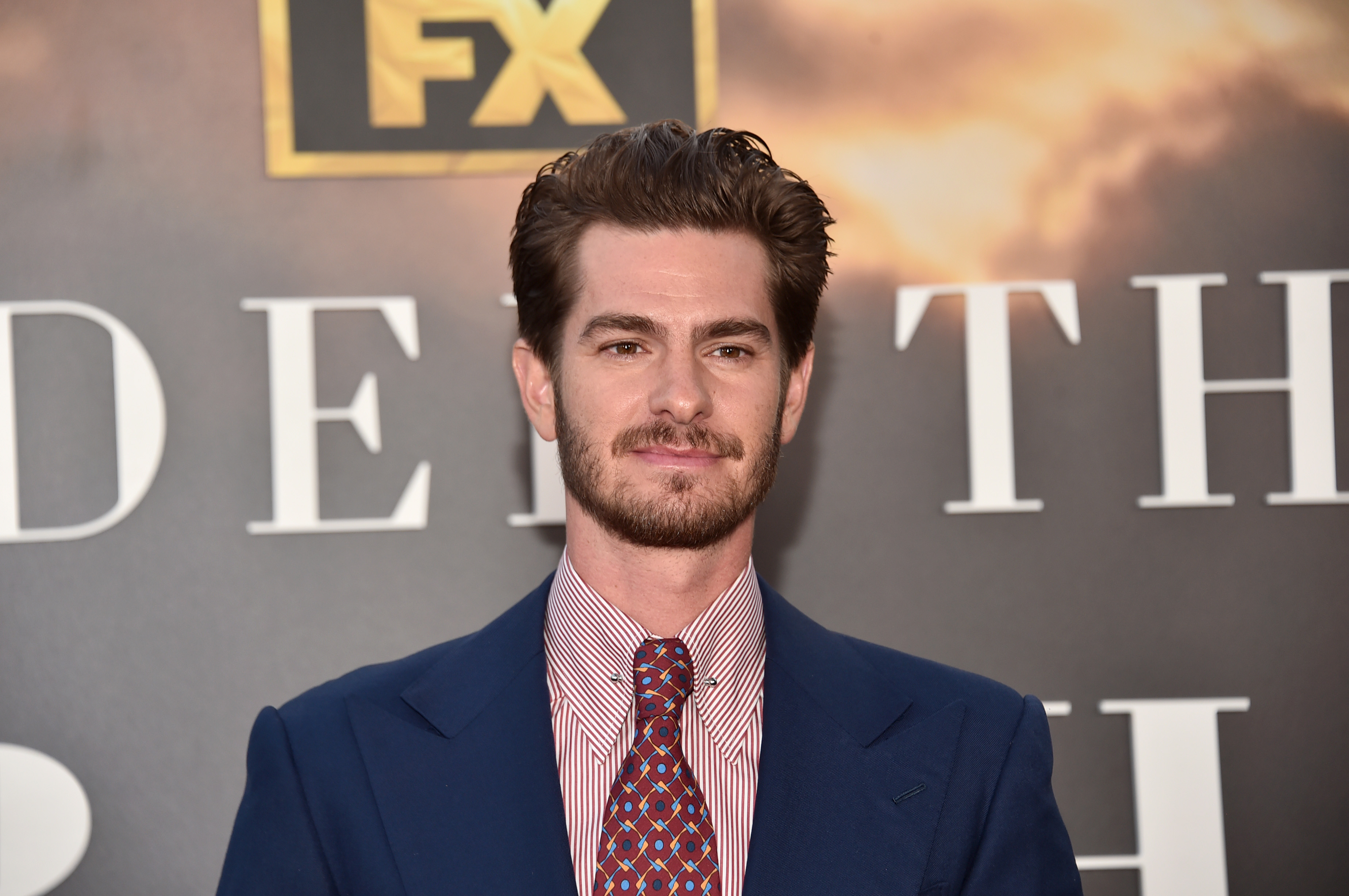 Andrew Garfield, who starred alongside Tobey Maguire in 'Spider-Man: No Way Home,' wears a dark blue suit over a light pink striped button-up shirt and dark red patterned tie.