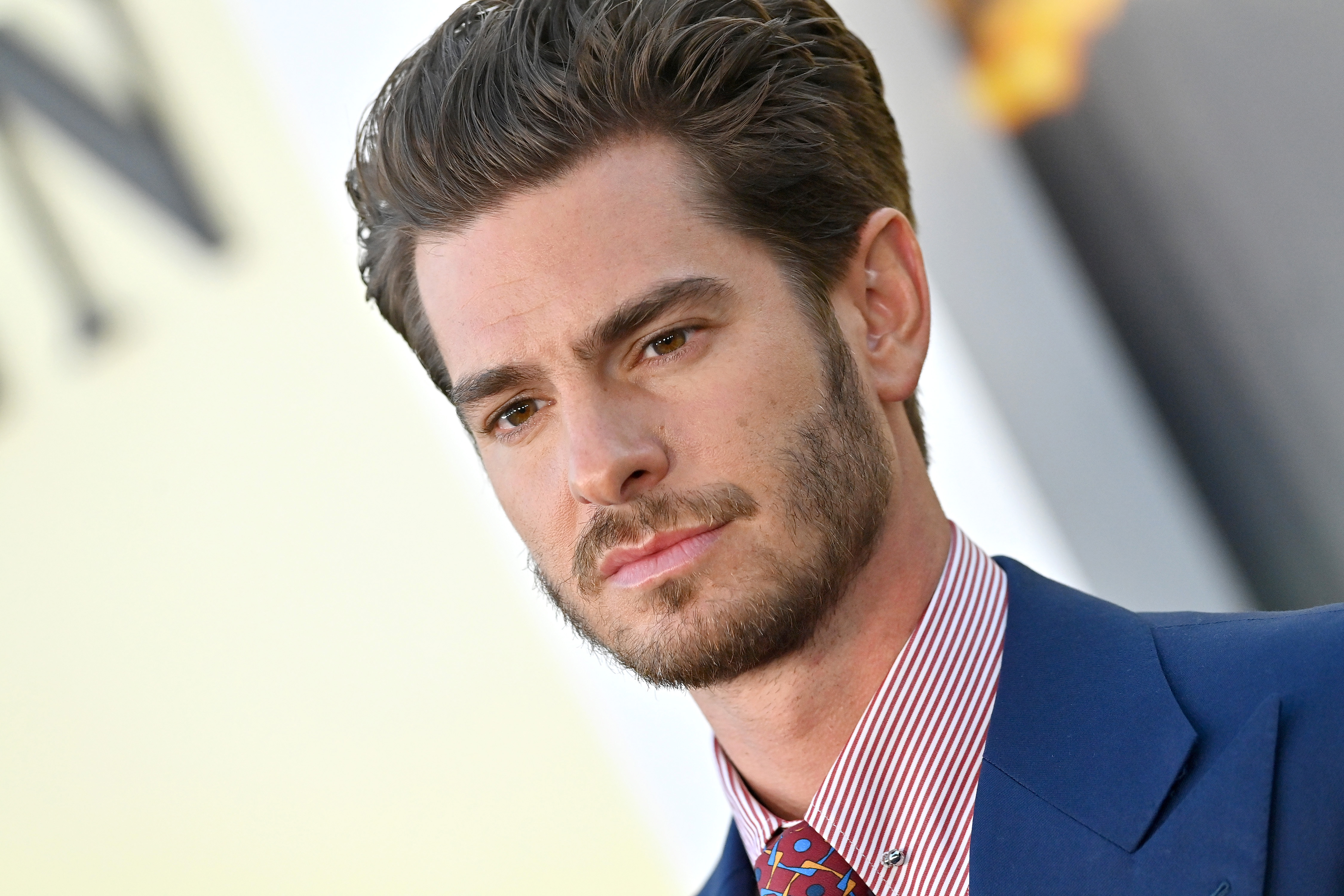 Andrew Garfield, star of 'Under the Banner of Heaven,' wears a blue suit over a red and white striped button-up shirt and red tie.