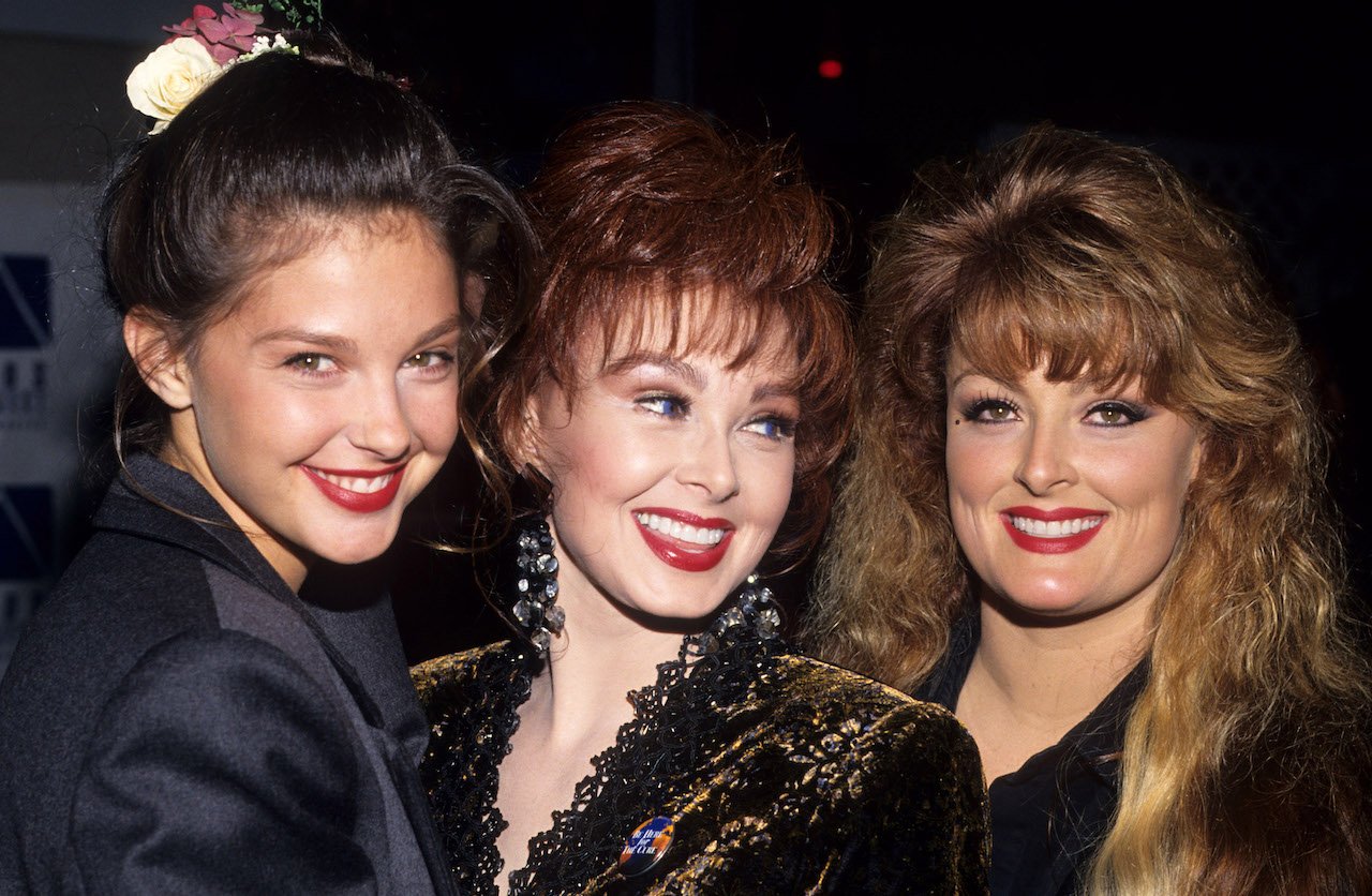 (L-R) Ashley, Naomi, and Wynonna Judd, all in black, posing in a huddle for a picture.