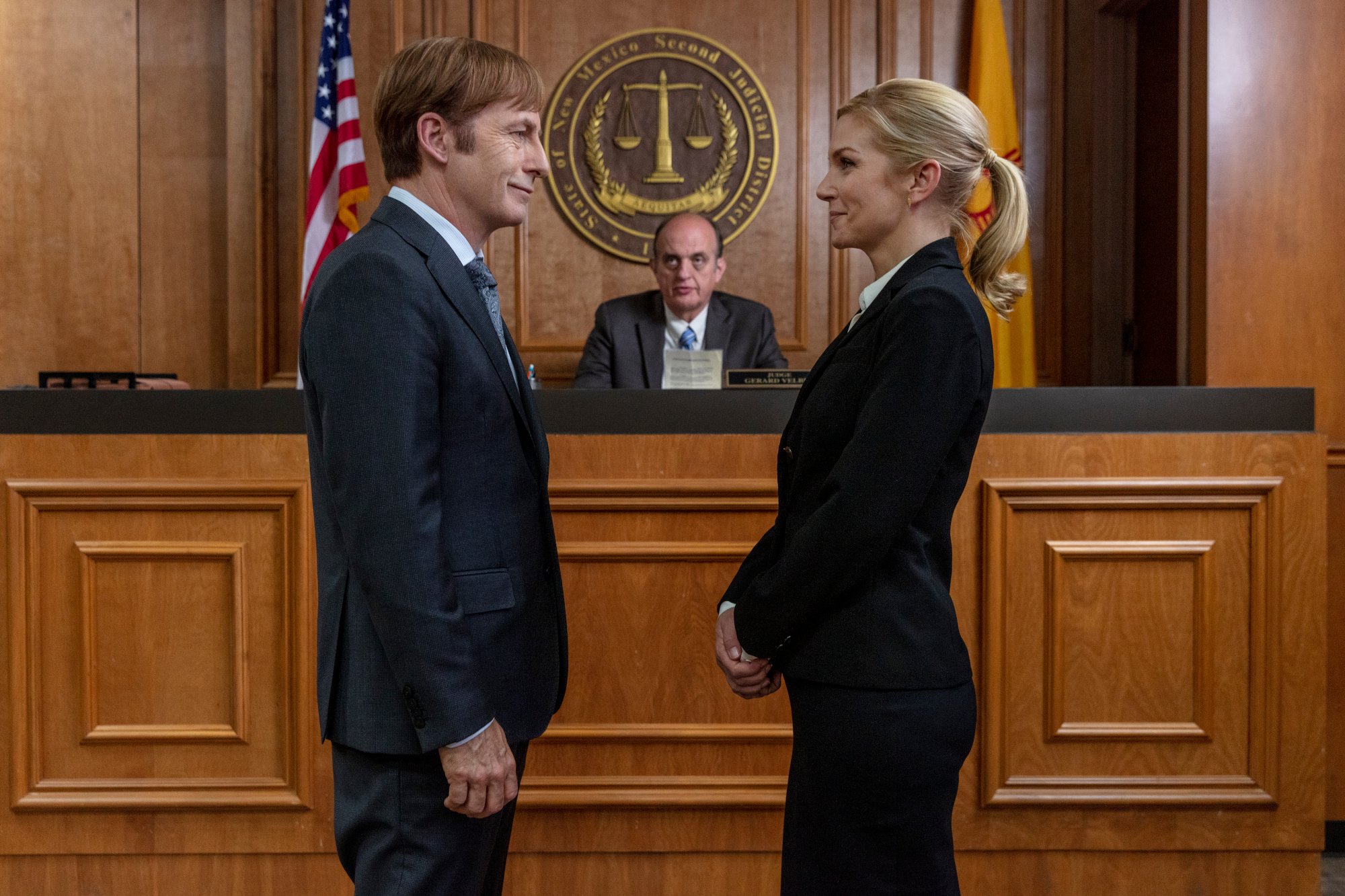 Bob Odenkirk and Rhea Seehorn as Jimmy McGill and Kim Wexler in 'Better Call Saul' Season 5. They're standing before a judge and getting married. They're both wearing dark suits and facing one another.