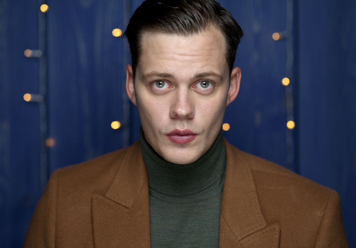Bill Skarsgård wears a brown jacket and looks directly into the camera.