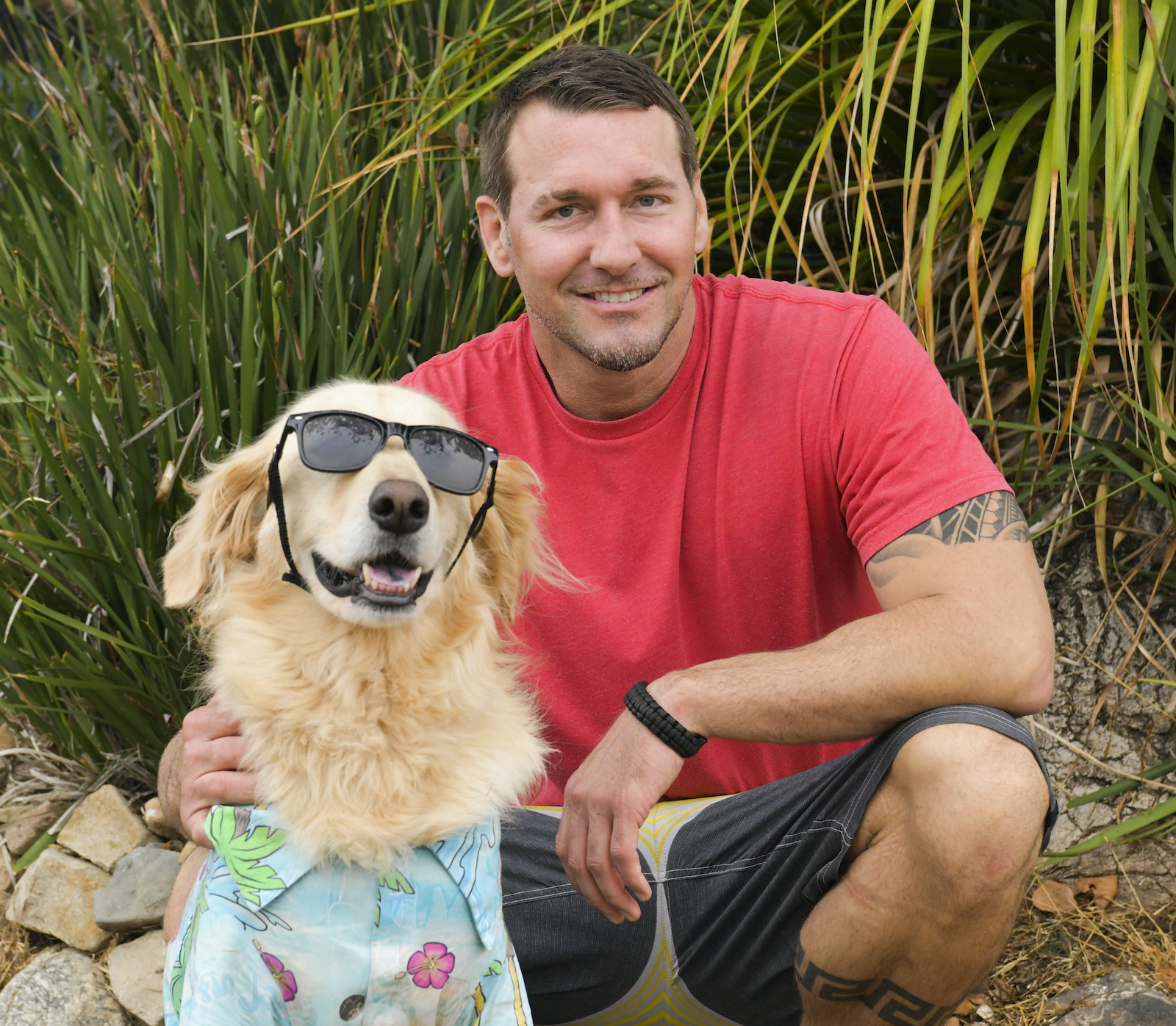 Dog trainer Brandon McMillan smiles with a dog who is wearing sunglasses and a shirt