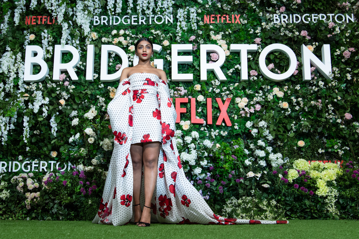 Charithra Chandran attends the Bridgerton Series 2 World Premiere at Tate Modern on March 22, 2022 in London, England. Chandran stands in front of a wall of flowers wearing a white dress with red flowers.