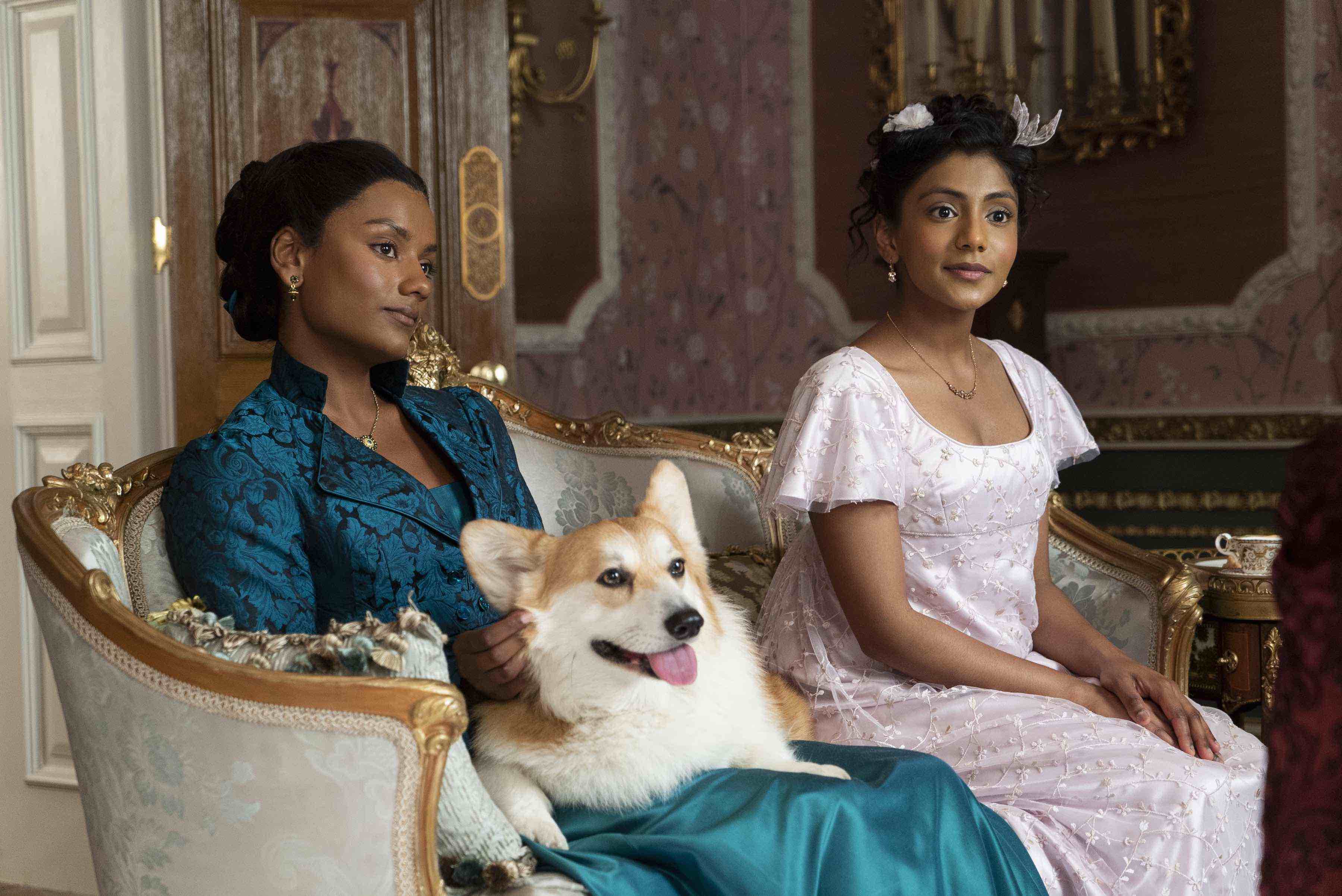 Simone Ashley and Charithra Chandran wear their "Bridgerton" costumes and sit on a fancy couch in a still from the show. 
