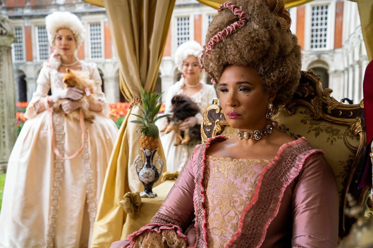 Queen Charlotte uses snuff in Bridgerton. In this photo she sits in a chair while women hold her dogs behind her.