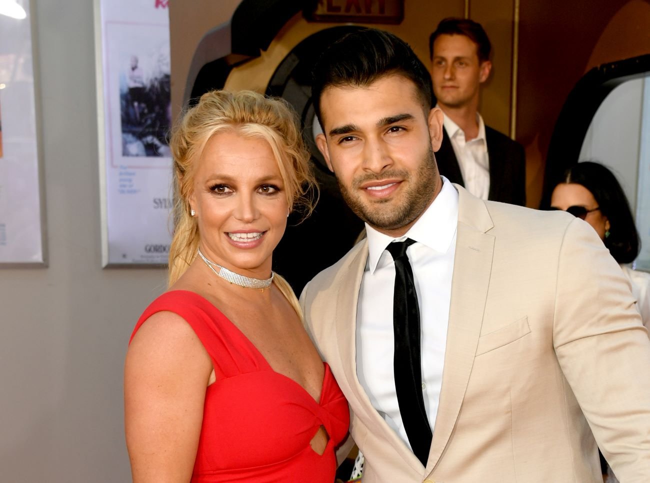 Britney Spears wears a red dress and stands next to Sam Asghari, who wears a tan suit.