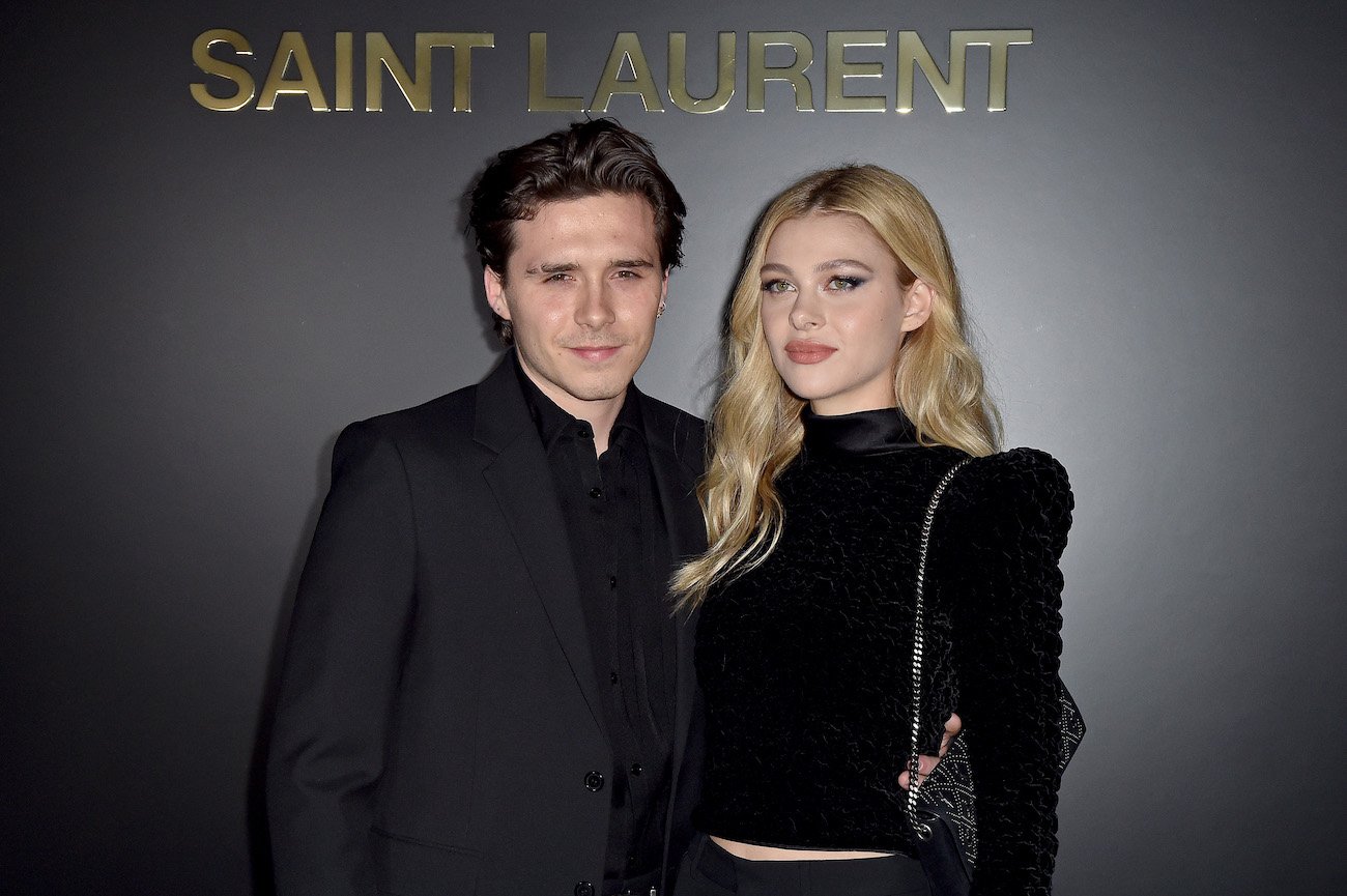 Brooklyn Beckham and Nicola Peltz both wearing black outfits and standing next to each other in front of a dark gray background