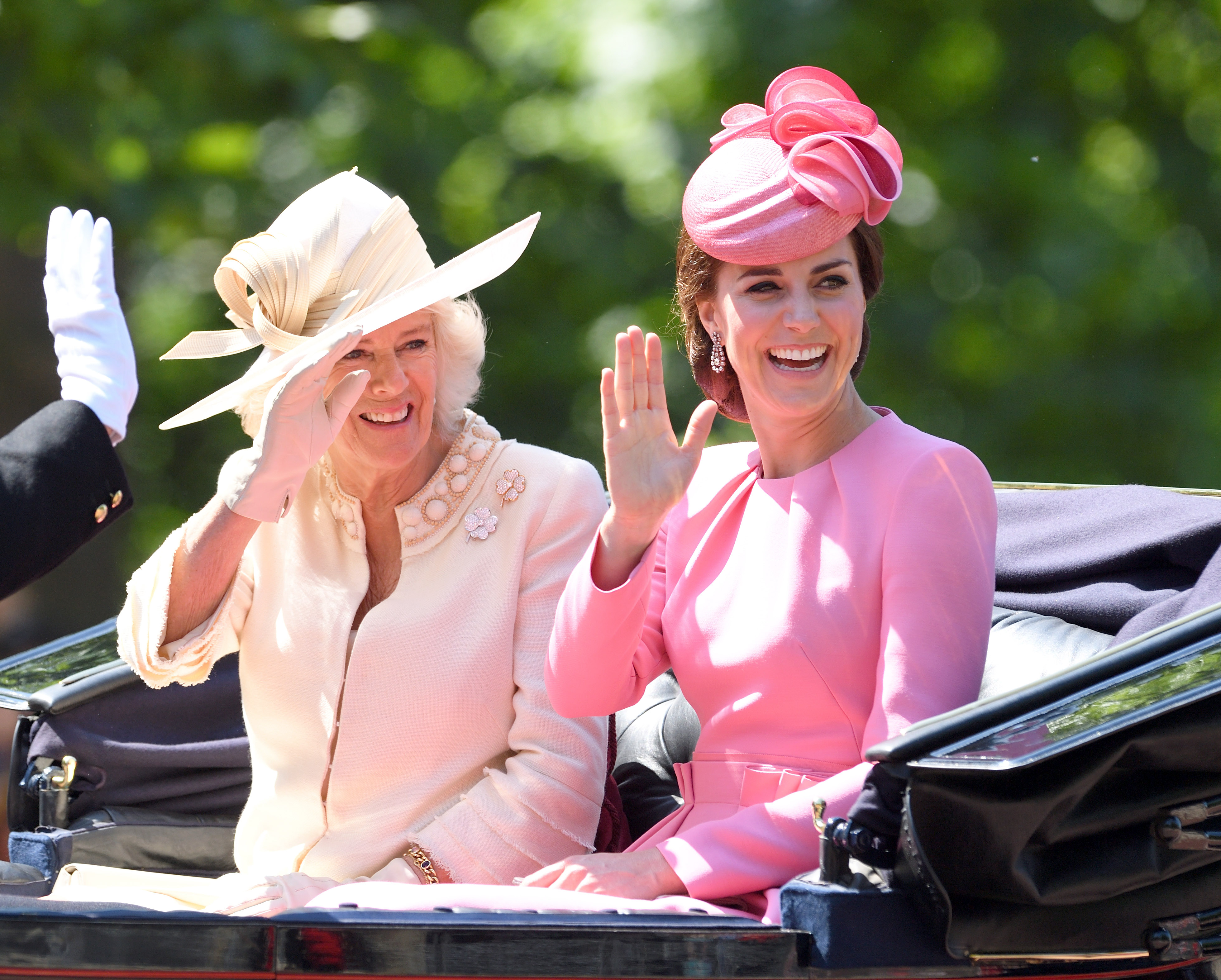 Camilla Parker Bowles wearing an off-white outfit with a fascinator and Kate Middleton wearing a pink outfit with a fascinator. Both women are waving while riding in a carriage