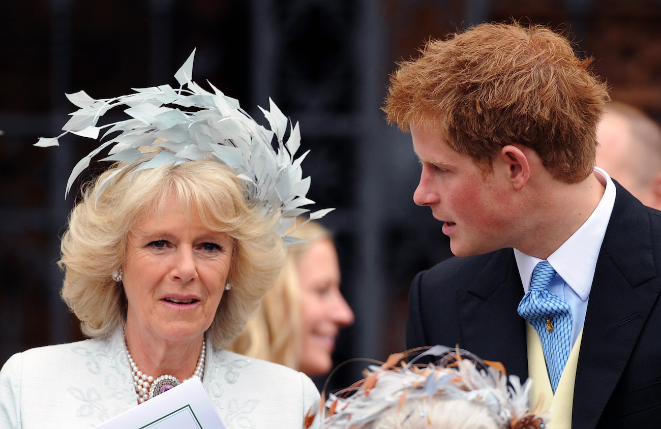 Camilla Parker Bowles looking on while wearing a fascinator. Prince Harry looking toward her while wearing a suit