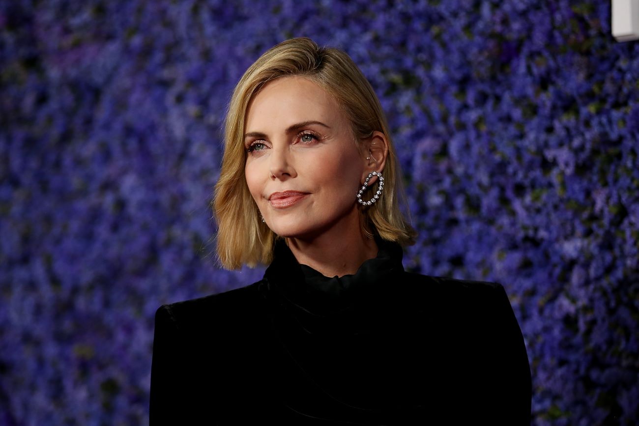 Charlize Theron wears black and stands against a dark blue background.