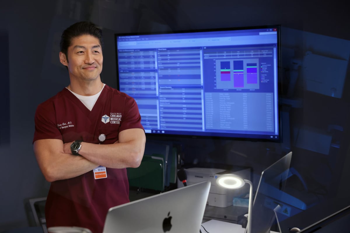 Brian Tee as Ethan Choi in Chicago Med Season 7. Choi folds his arms and smiles. He is standing in front of a screen.