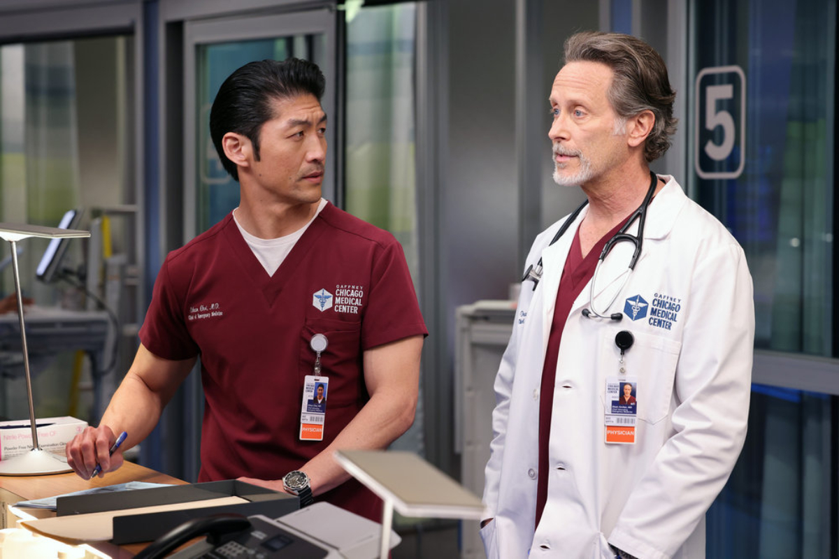 Brian Tree as Ethan Choi and Steven Weber as Dr. Dean Archer in Chicago Med Season 7. Ethan and Archer stand at a desk. 