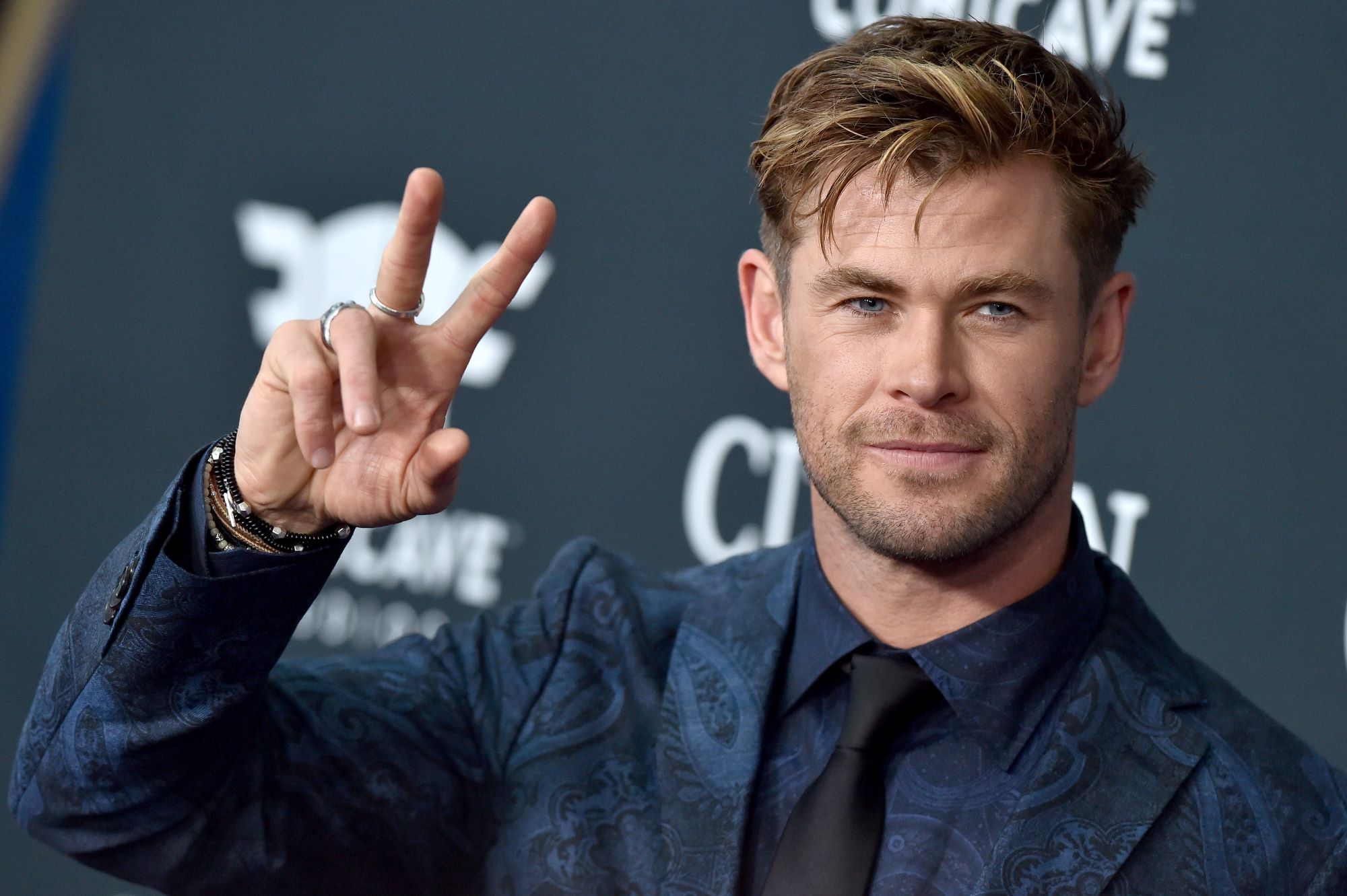 Chris Hemsworth, star of 'Thor: Love and Thunder,' which still hasn't released its trailer, holds up a peace sign while wearing a dark blue paisley suit and black tie.