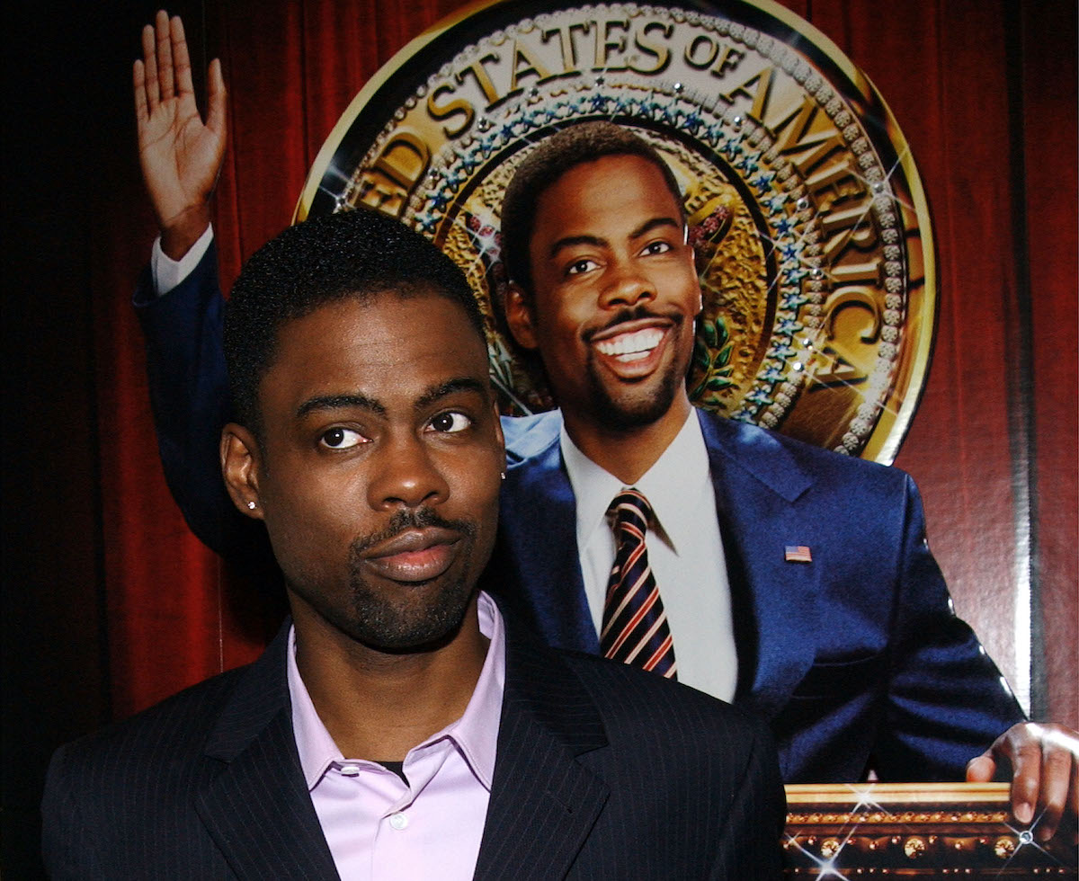 Chris Rock wears a suit and poses in front of a poster for ‘Head of State’