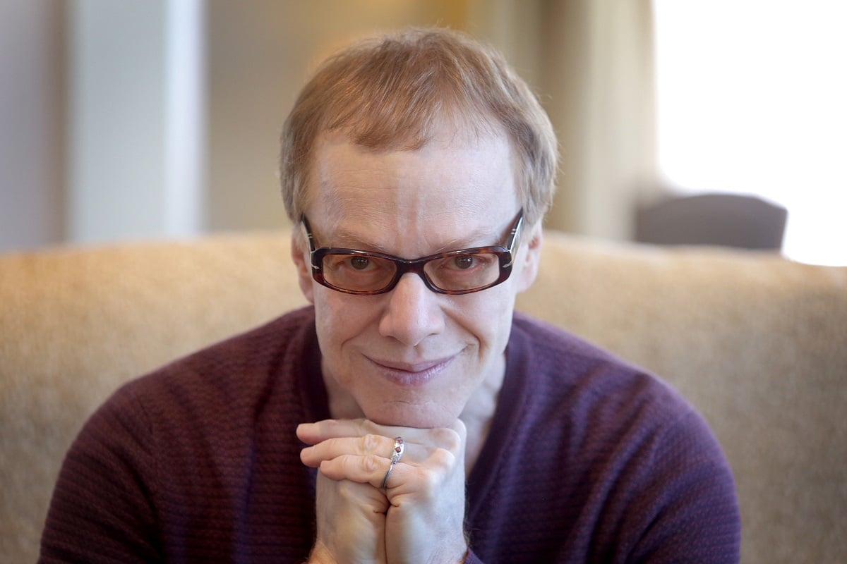 Danny Elfman sitting down smirking while wearing glasses.