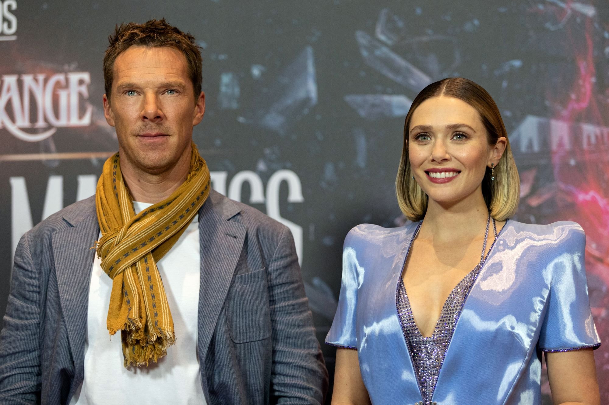 Benedict Cumberbatch and Elizabeth Olsen, who star as Doctor Strange and Scarlet Witch in 'Doctor Strange 2,' pose for pictures together. Cumberbatch wears a gray suit over a white shirt and yellow scarf. Olsen wears a short-sleeved light blue jacket over a bedazzled silver shirt.