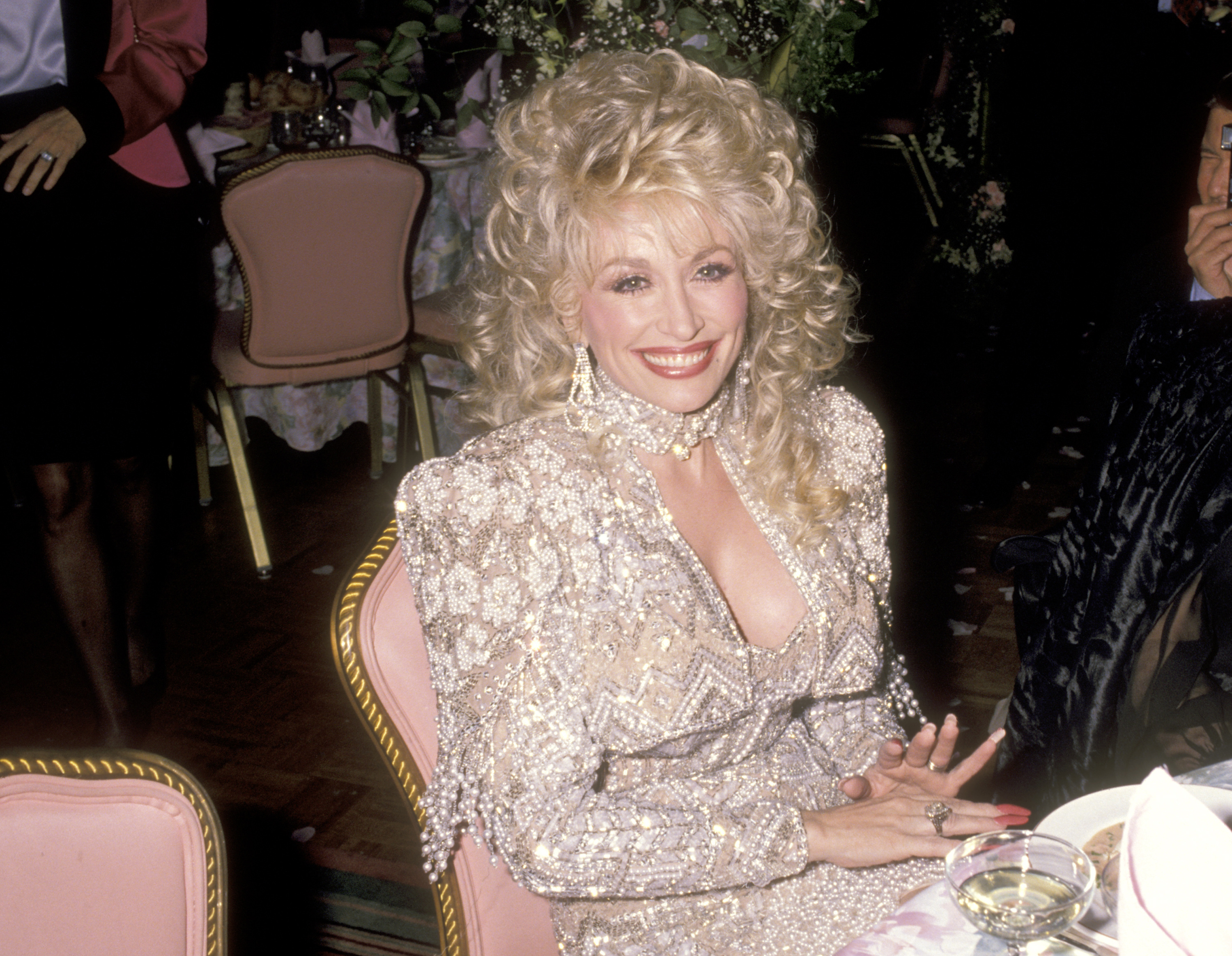 Dolly Parton wears a white beaded dress and sits in a pink chair at the 'Steel Magnolias' premiere.